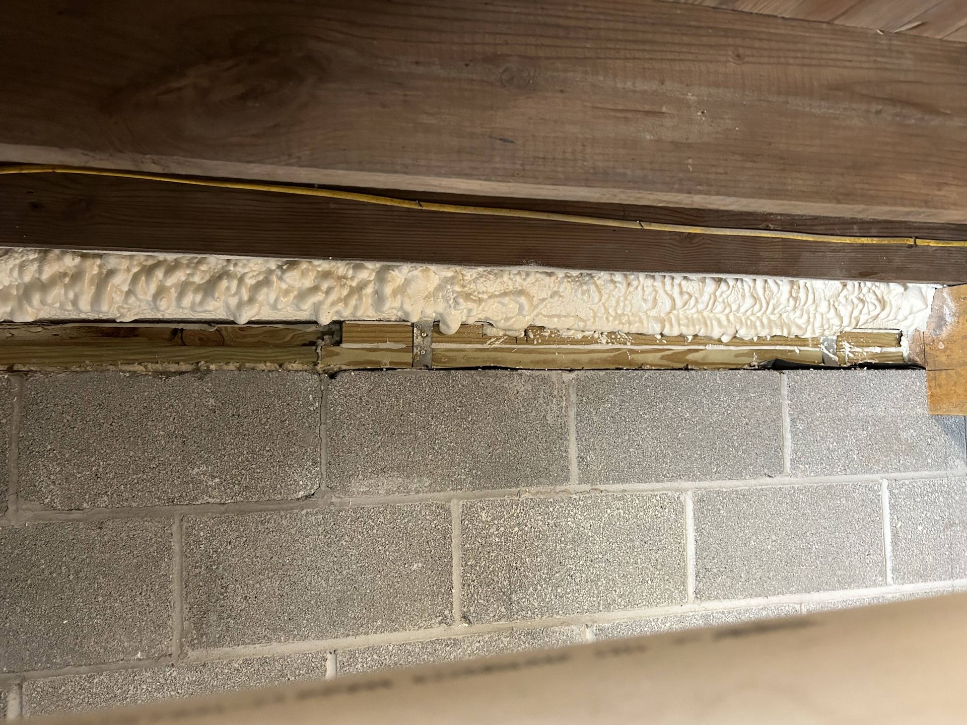 Close-up of exposed foam insulation between a wooden beam and a gray cinder block wall