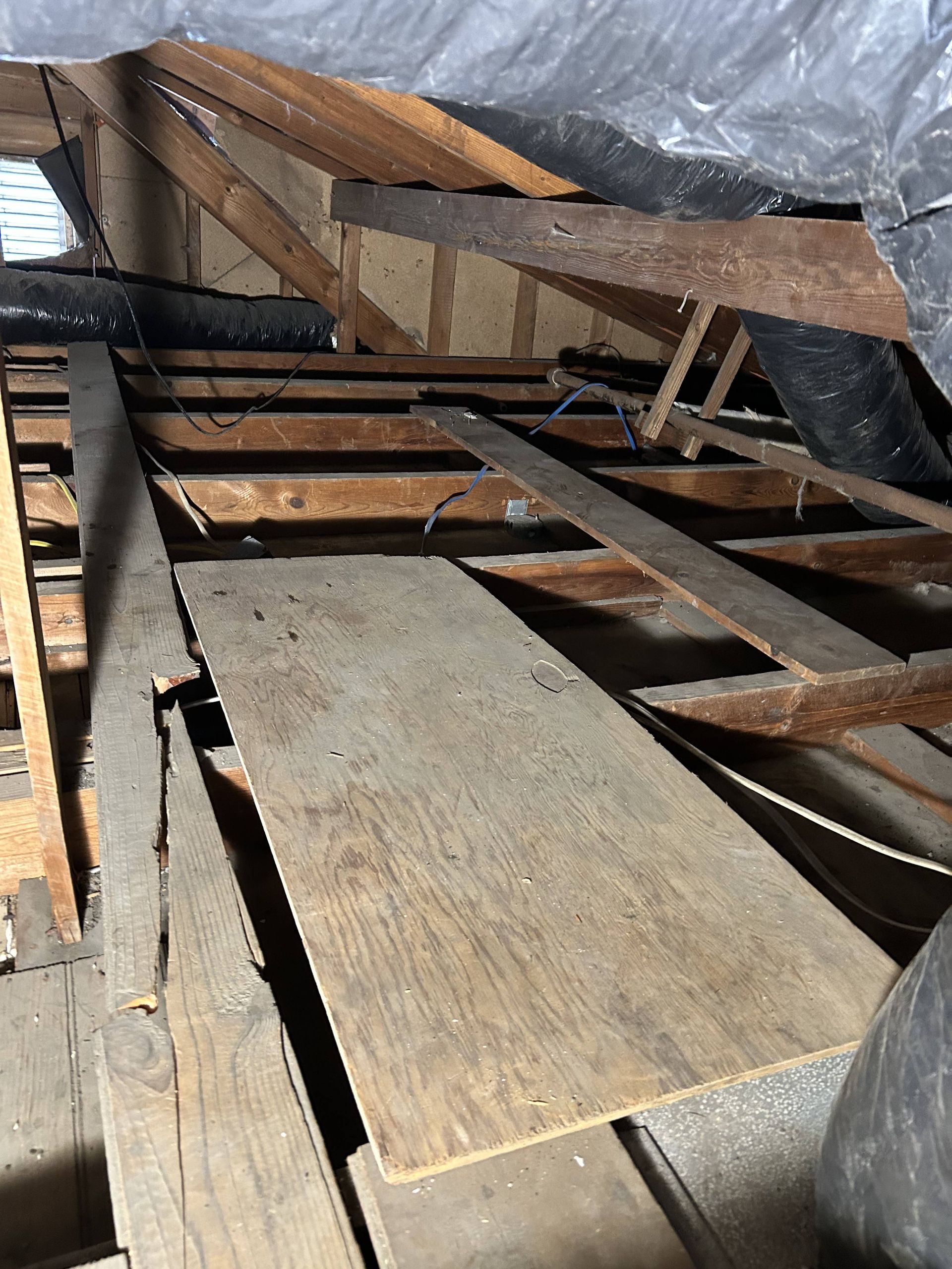 Damaged wooden floor joists and planks in a collapsed attic or crawl space, with debris and plastic sheeting