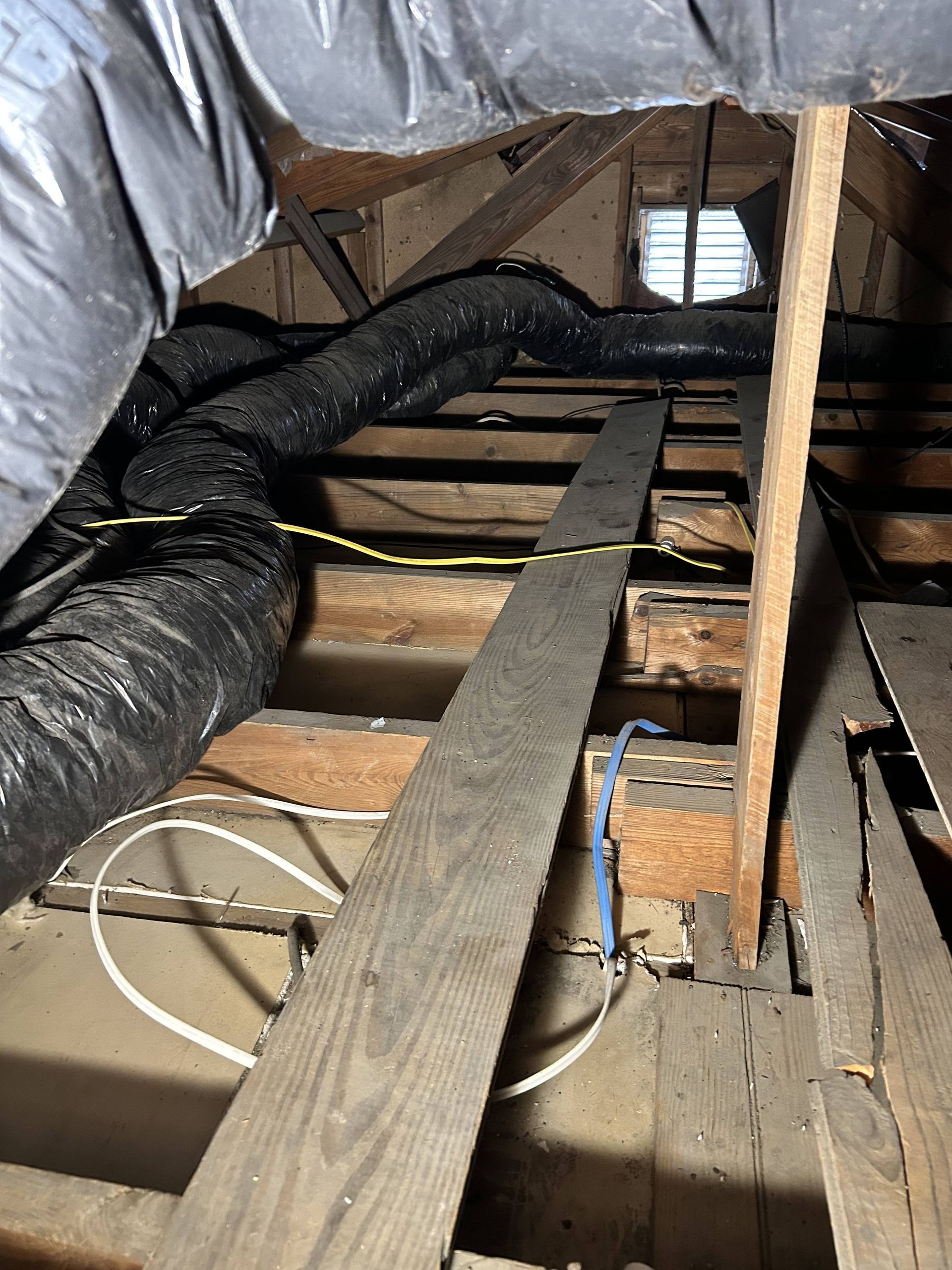 Attic crawlspace with exposed wooden joists, insulation, and black ducting, viewed along a narrow plank walkway