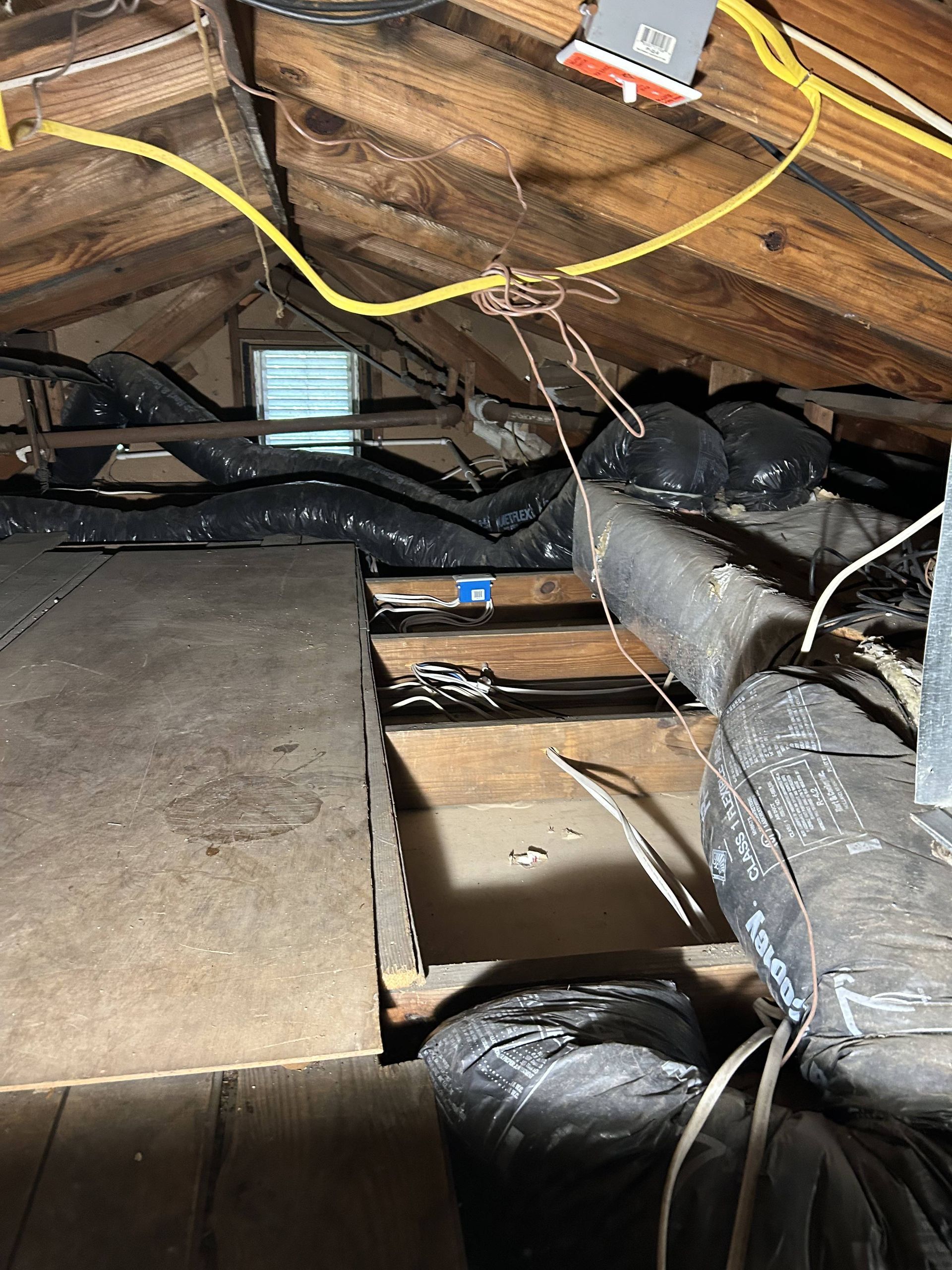 Attic with exposed rafters, insulation, and hanging cables around a duct and narrow walkway