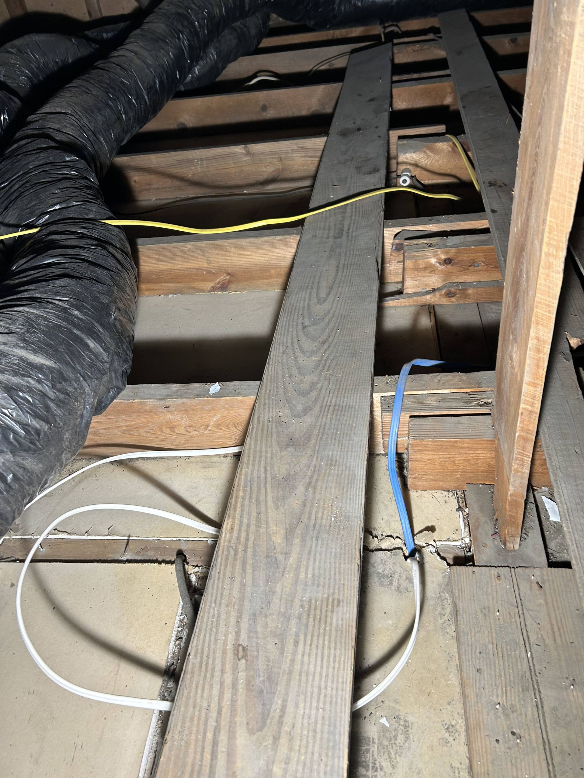 Exposed attic floor joists with black ducting and loose electrical cables.