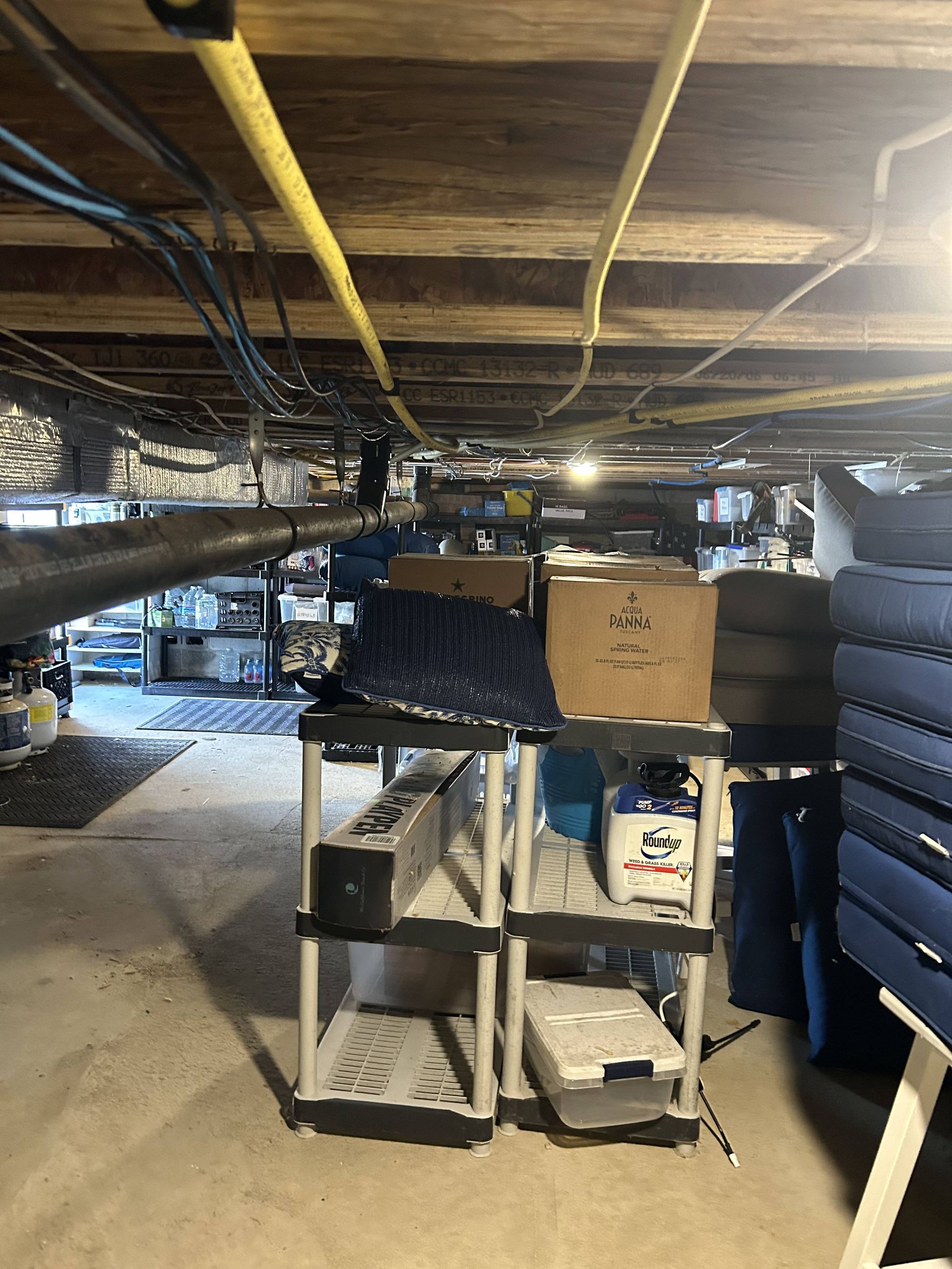 Cluttered basement under wooden beams with shelving, boxes, and stacked tires beside a concrete floor