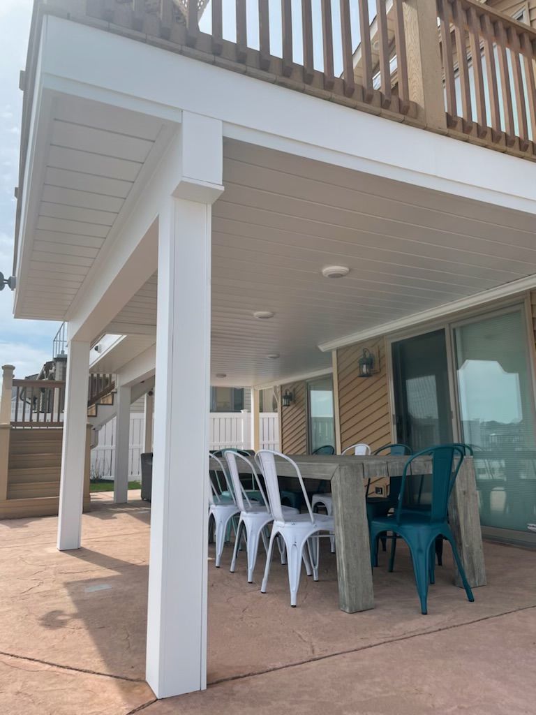 Covered patio with dining table and chairs beneath an elevated deck, beside a house with sliding glass doors.
