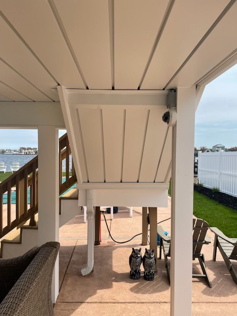 White outdoor shower enclosure under a porch, with a hose, steps, and beach view beyond the deck.