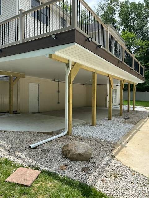 Covered driveway under a raised building, with gravel, support posts, and a concrete walkway.