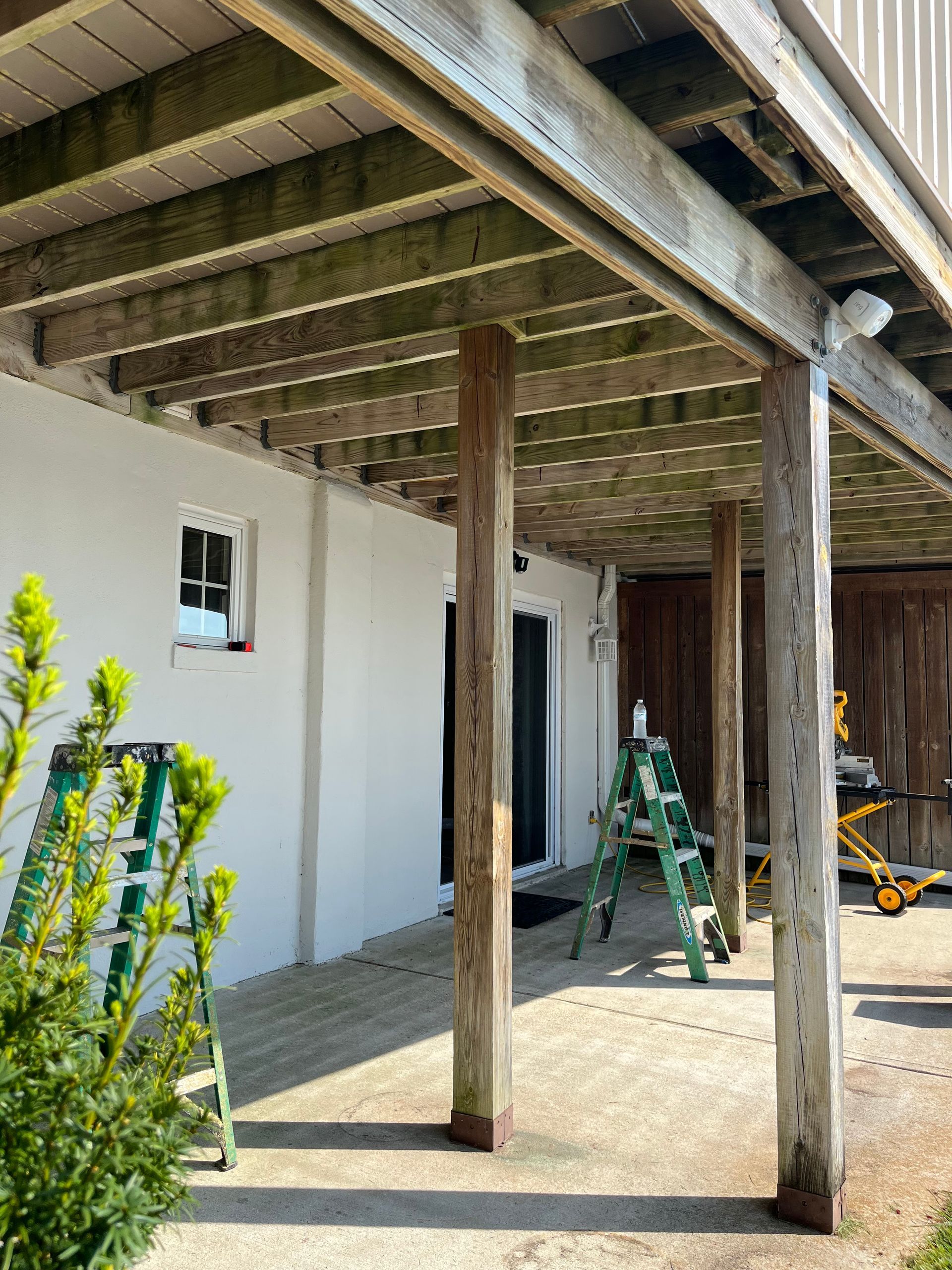 Wooden porch under construction beside a white house, with ladders and tools in a shaded patio area