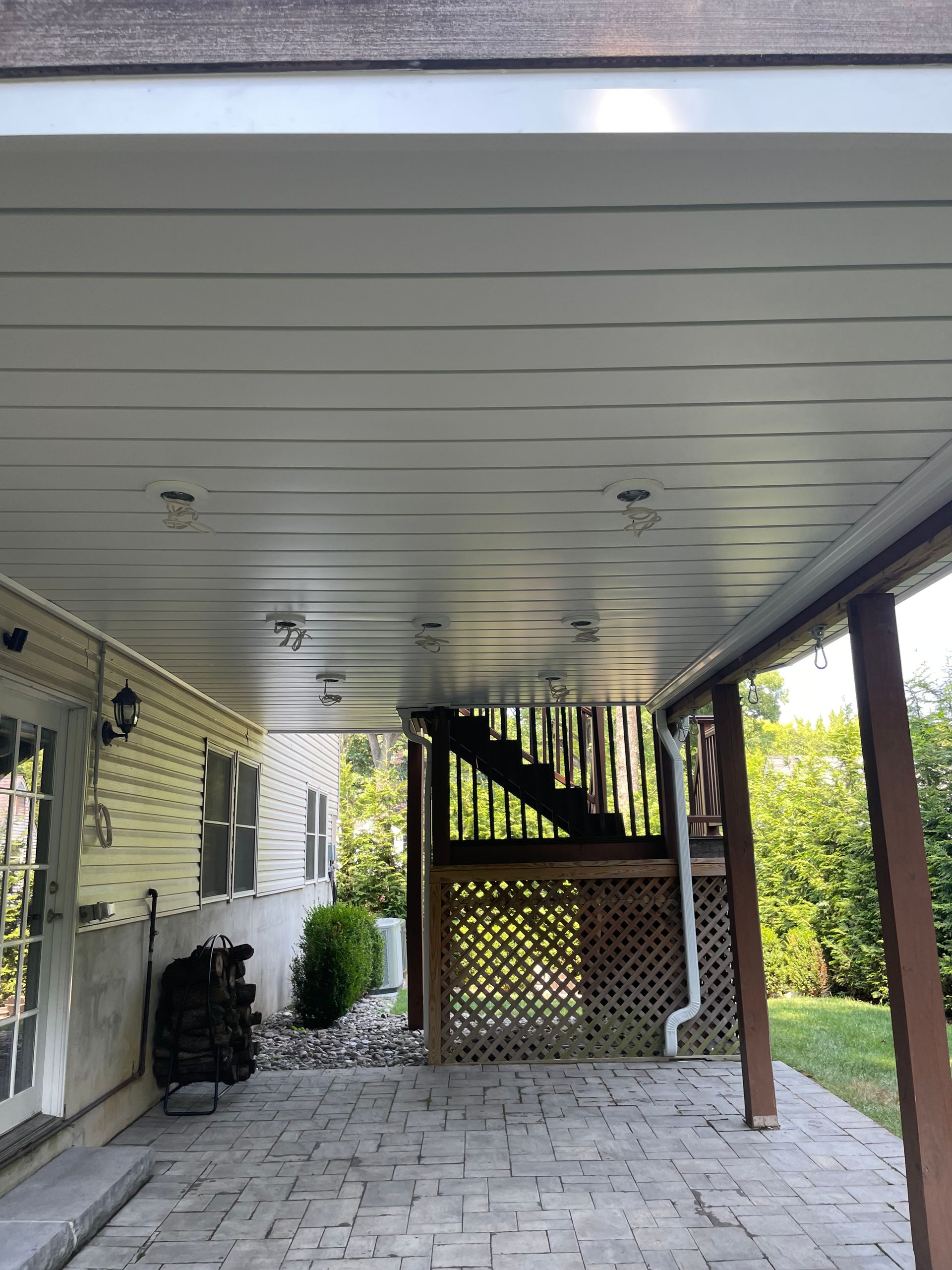 Covered patio with gray ceiling, stone floor, wooden stairs, and lattice fence beside a house