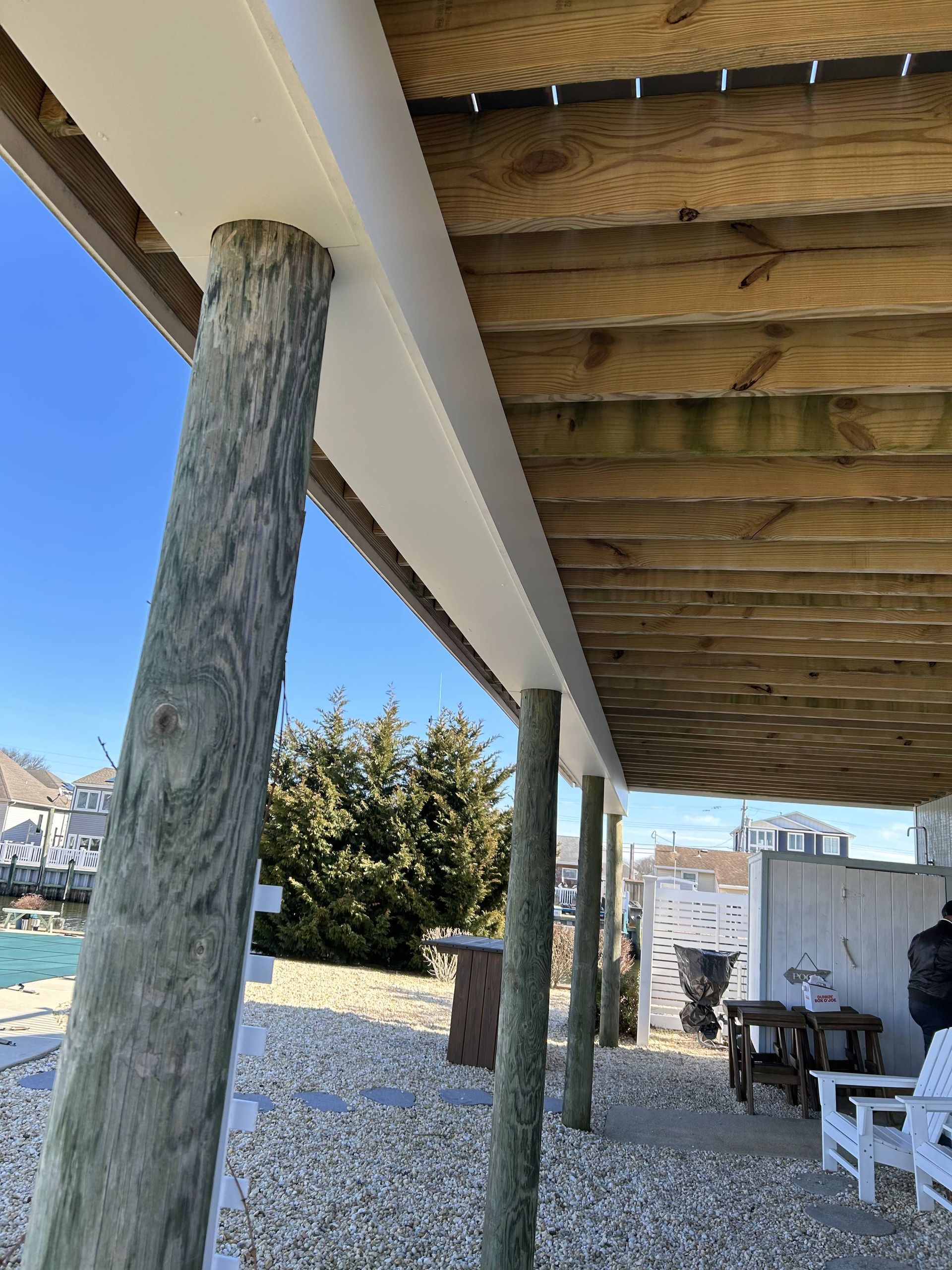 Underside of a porch with wooden beams, log posts, and white chairs beside a gravel yard