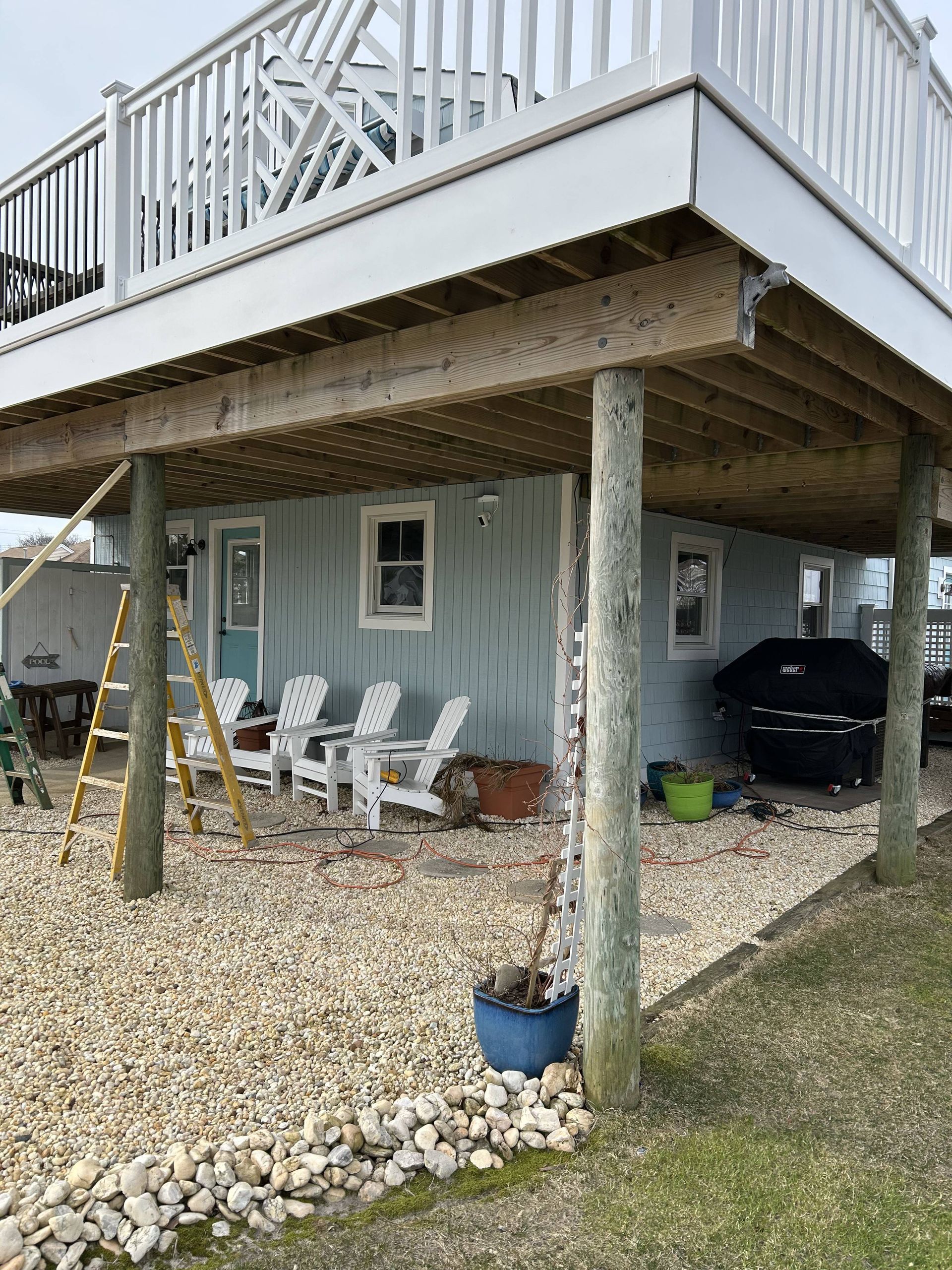 House under raised deck with white chairs, gravel yard, and a black grill on the patio.