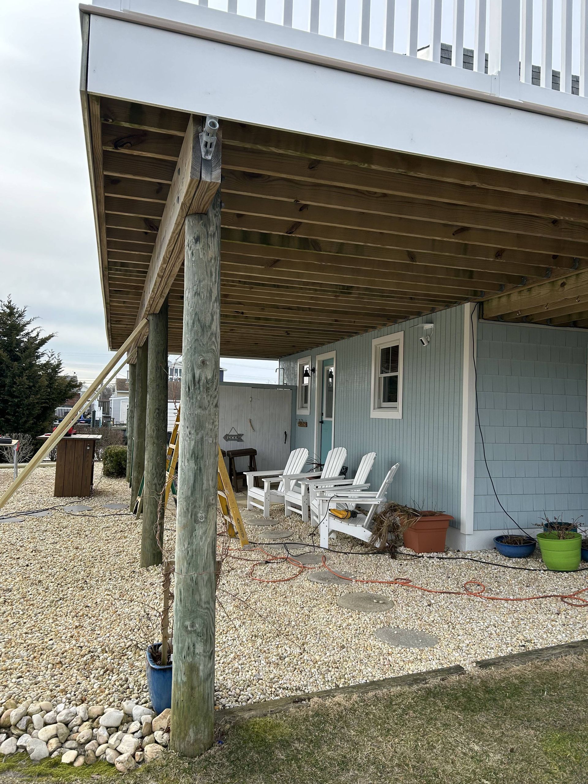 House under raised deck with gravel yard, white chairs, and potted plants.