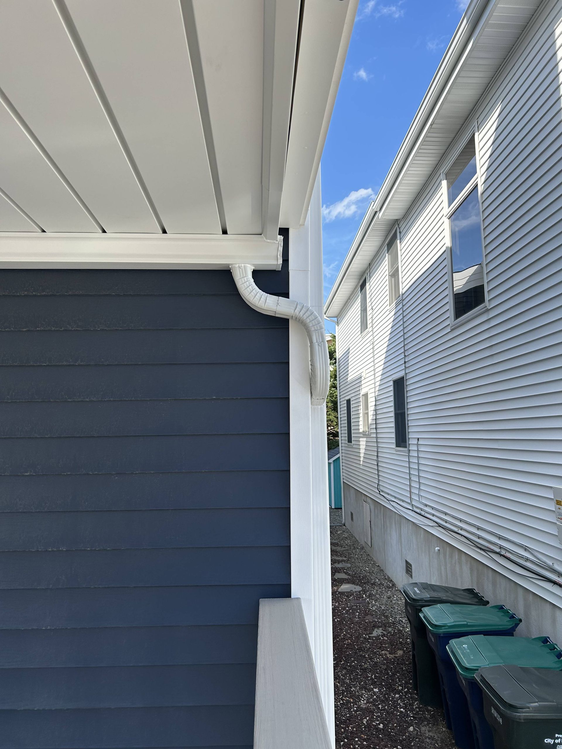 Narrow side yard between two houses, with blue siding, white trim, and trash bins along a fence.