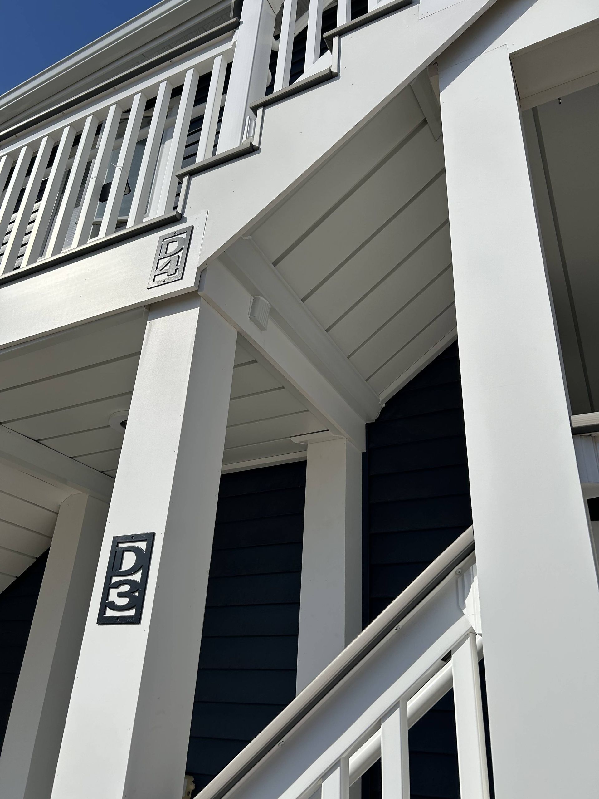 White porch stairway and railings on a house exterior, with dark siding and a number plaque by the column.
