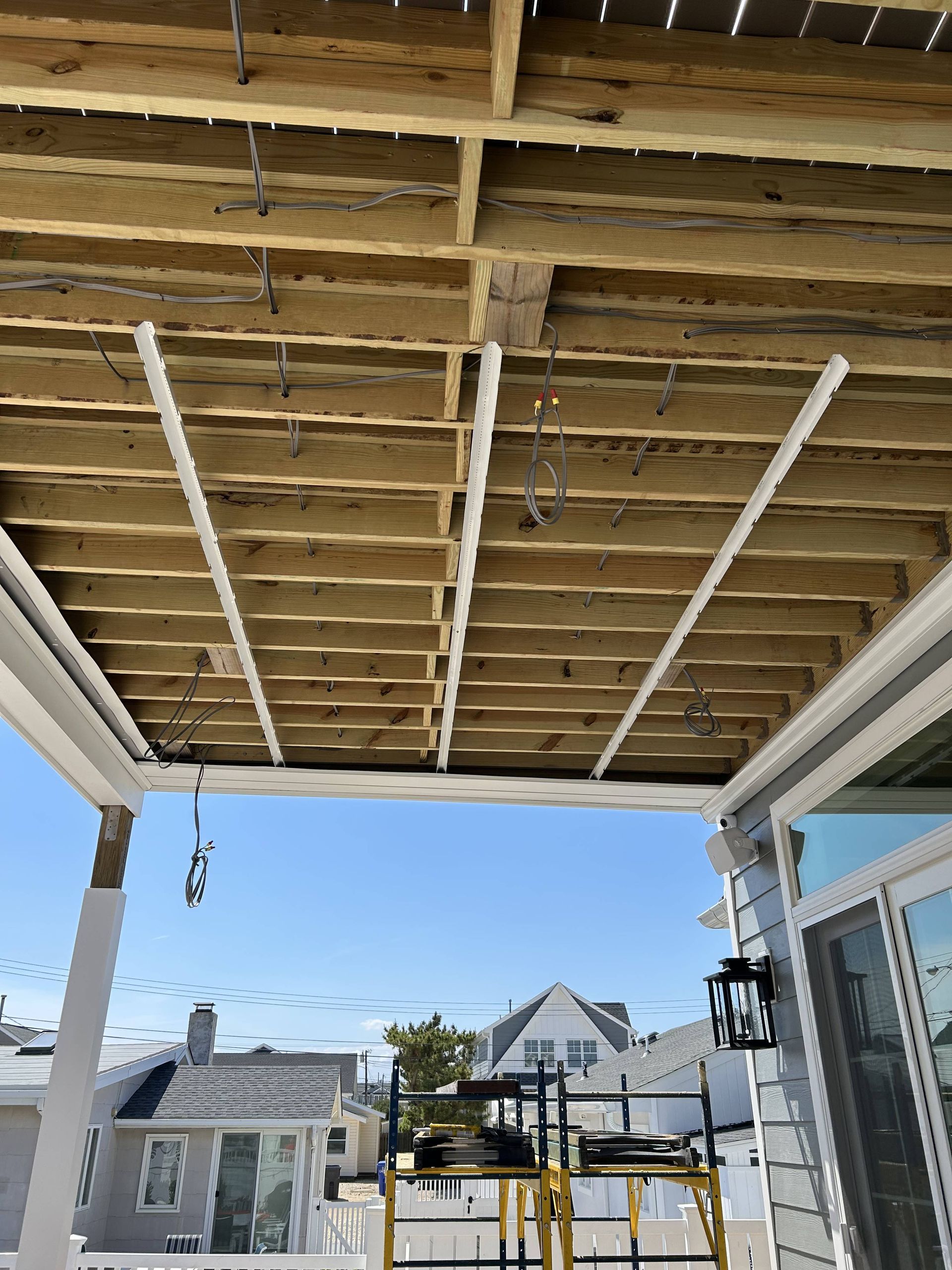 Under-construction porch ceiling with exposed wooden joists and hanging white support beams overlooking houses outside