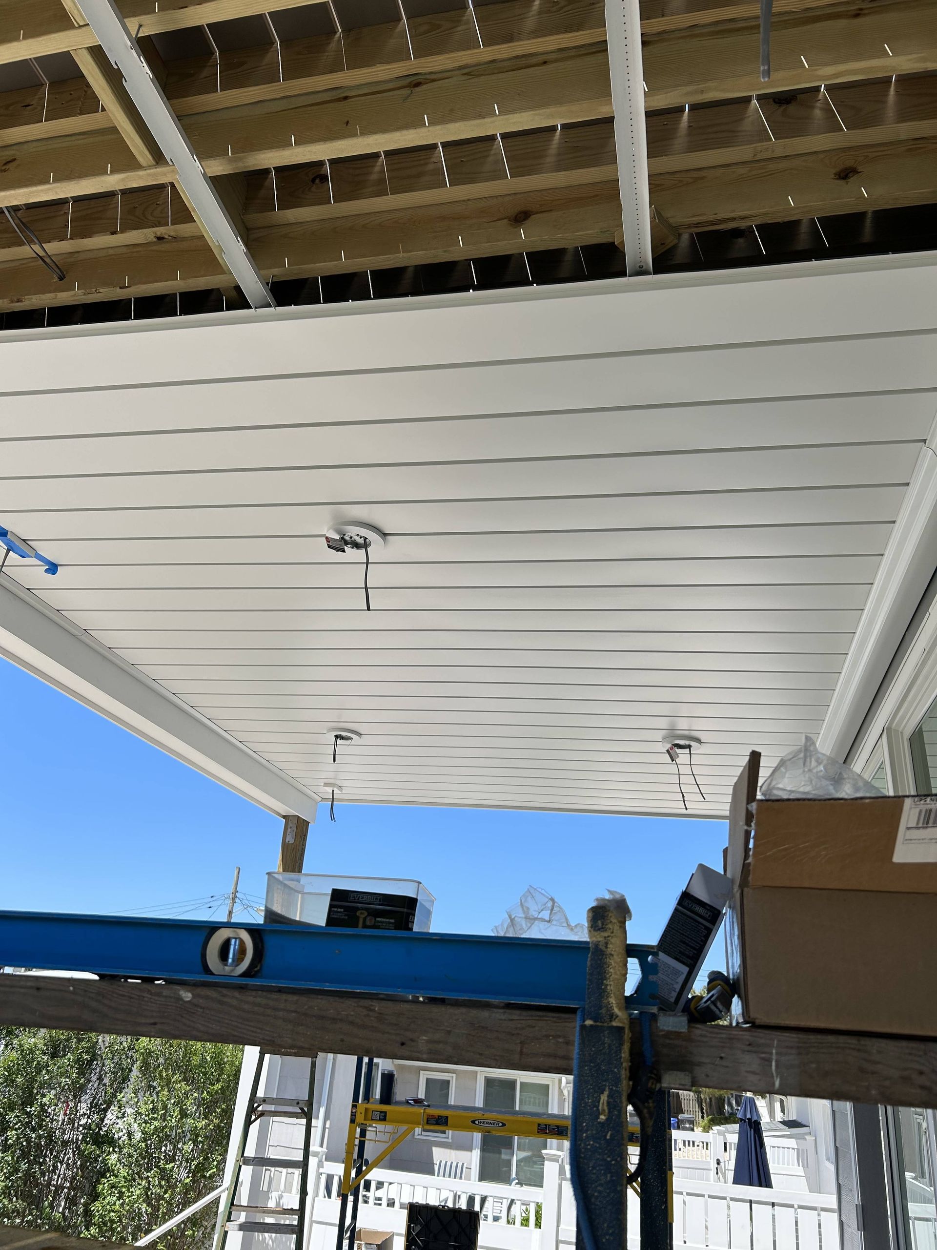 White slatted patio ceiling under construction, with blue scaffolding and exposed wooden beams above.