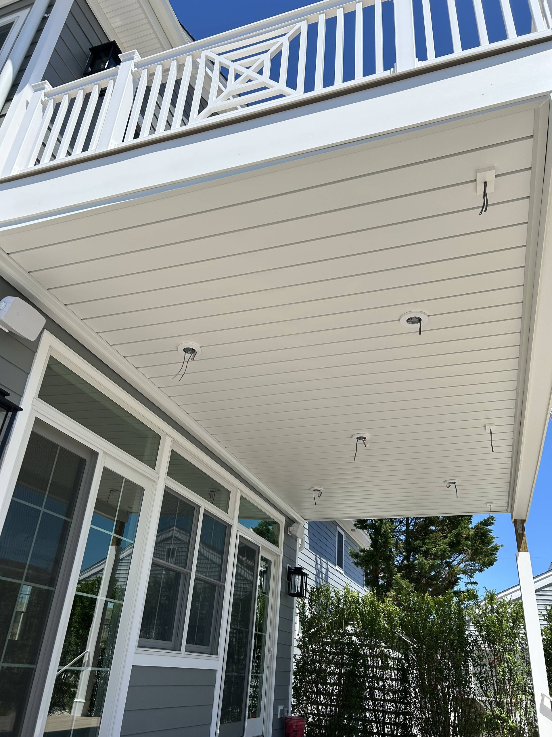 White porch ceiling with recessed lights beneath a balcony, beside glass doors and windows.