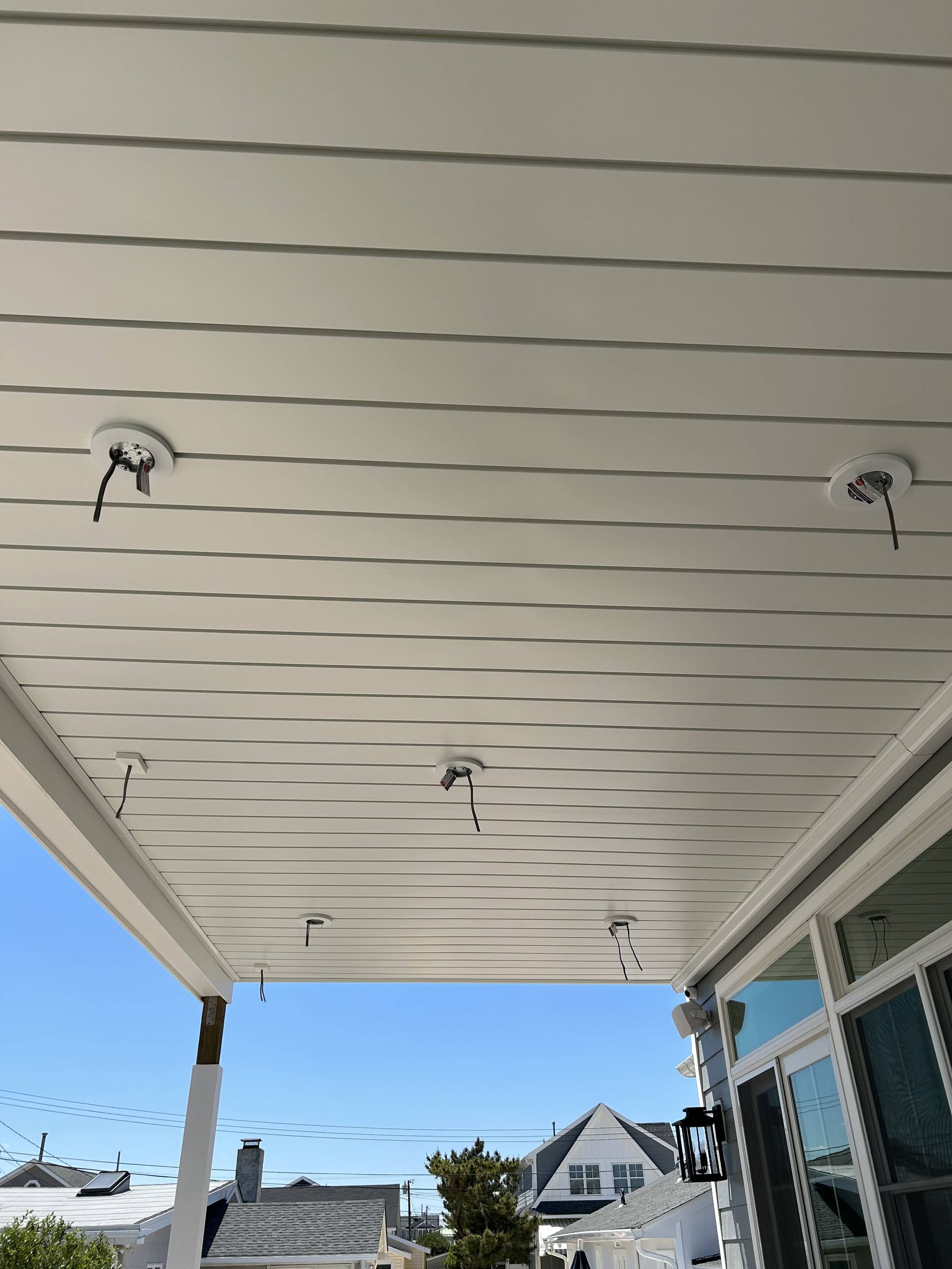 View from a porch ceiling with white beams and lights, overlooking neighboring houses and blue sky.