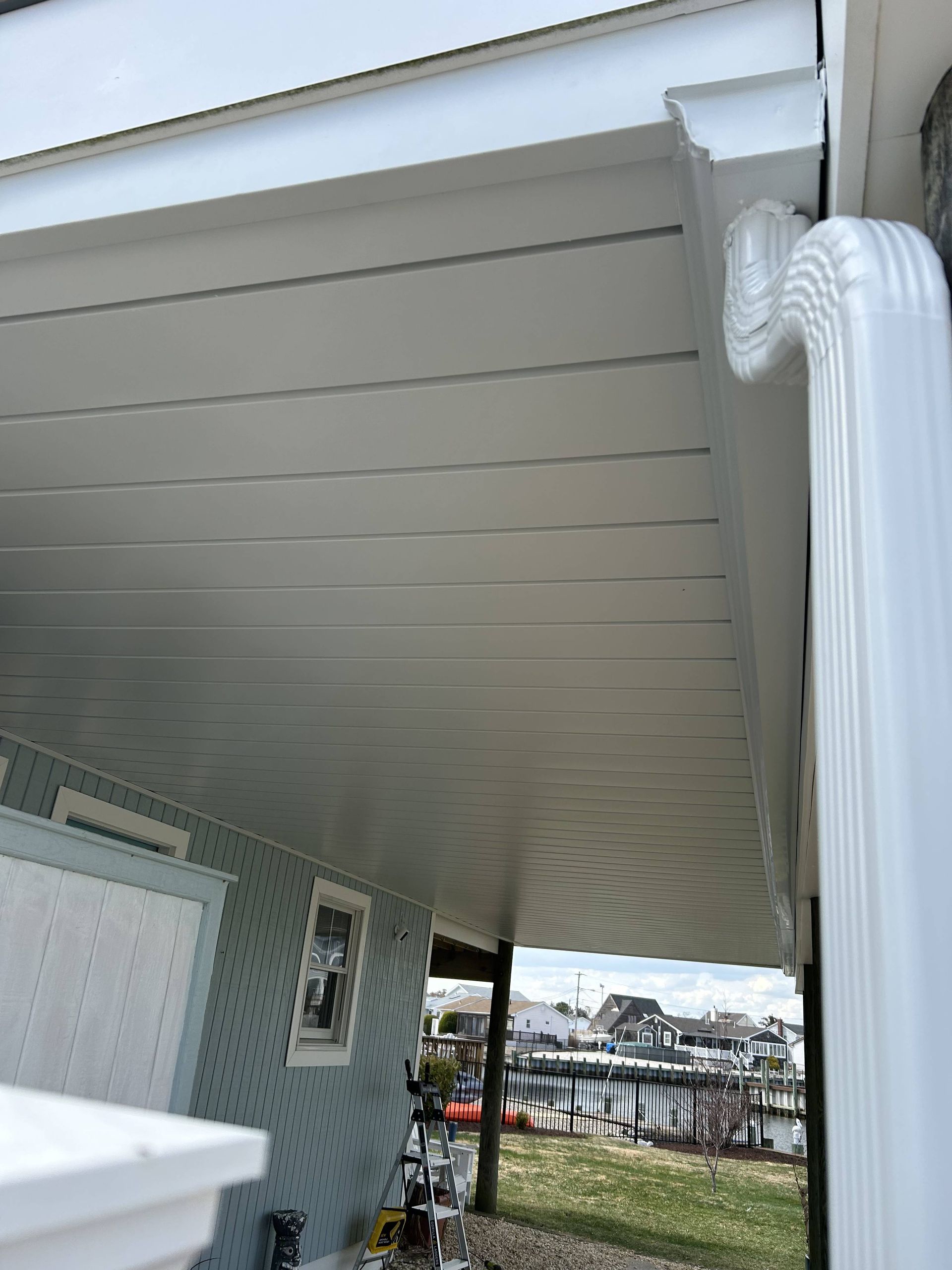 View under a house porch, showing white siding, ceiling panels, and a glimpse of water and boats beyond.