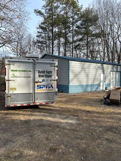 White enclosed trailer parked beside a gray metal building in a wooded yard.