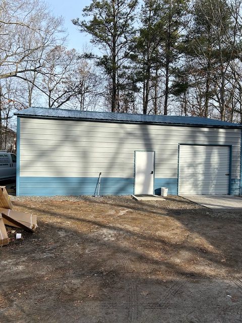 White metal garage or shed with blue trim, two doors, and trees in the background on a dirt lot.