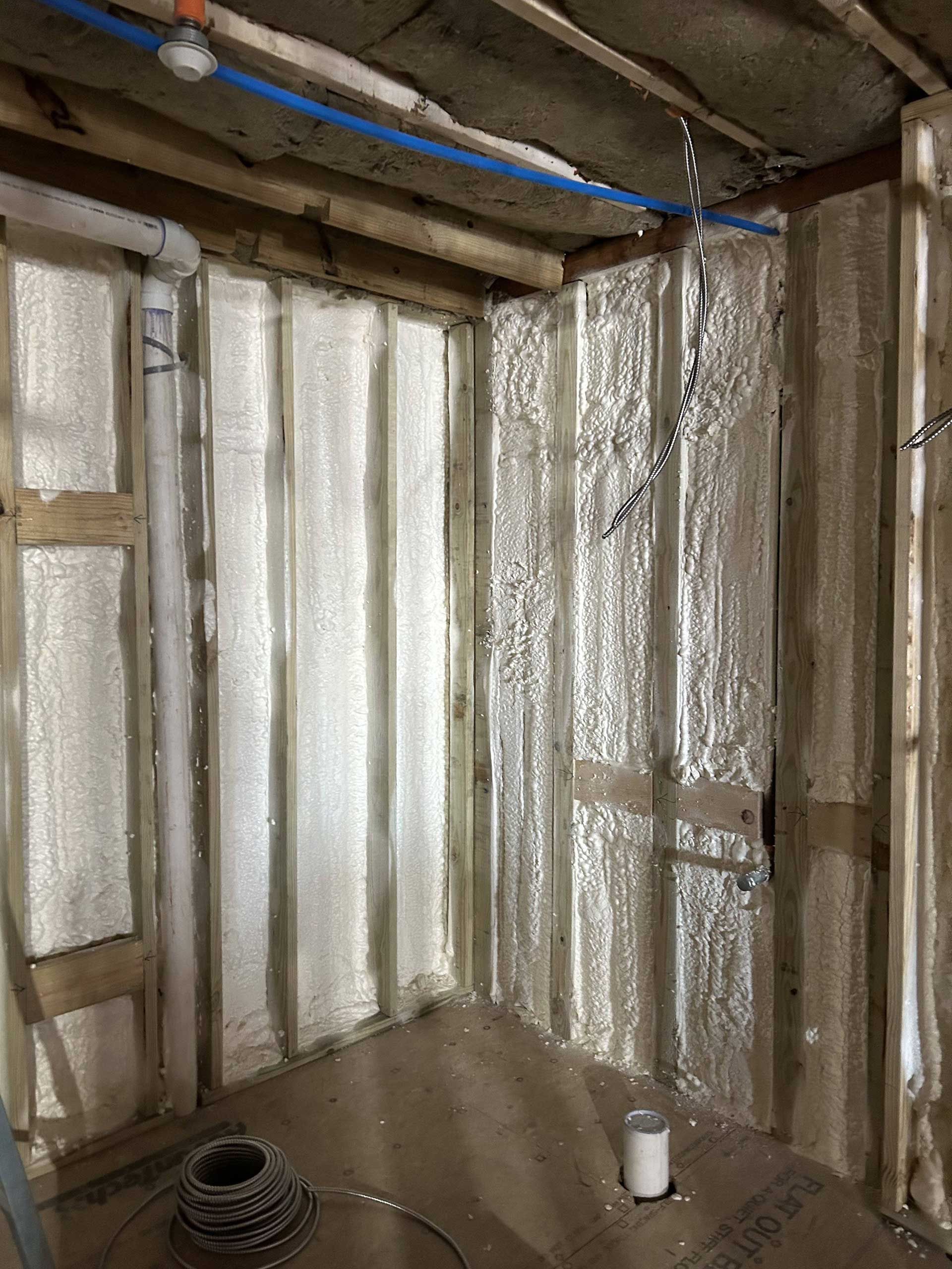 An unfinished interior corner room showing wall studs insulated with spray foam, a plumbing pipe, and exposed wiring.