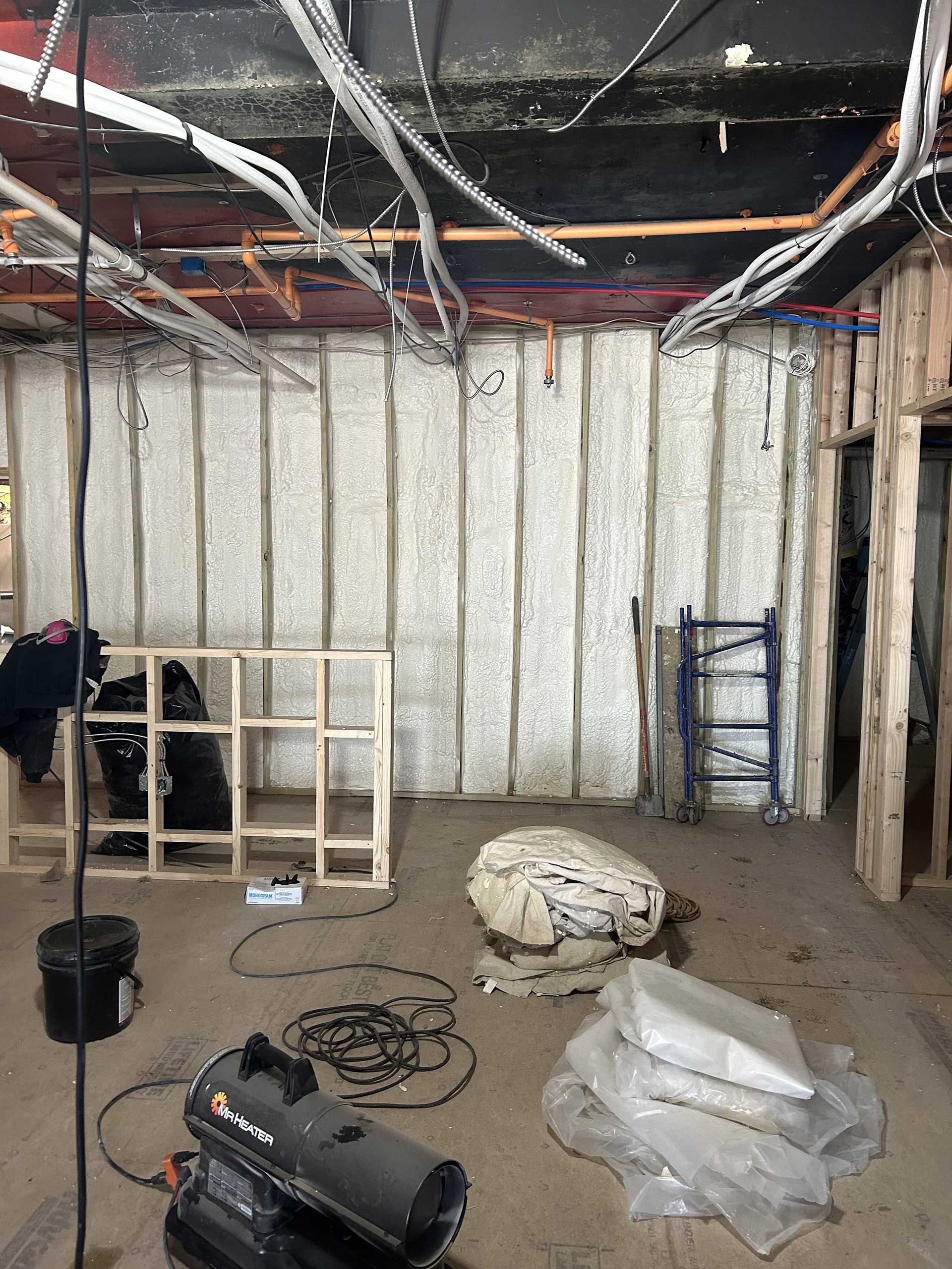 Interior construction site with exposed wooden studs, foam-insulated walls, ceiling wiring, and scattered building debris.