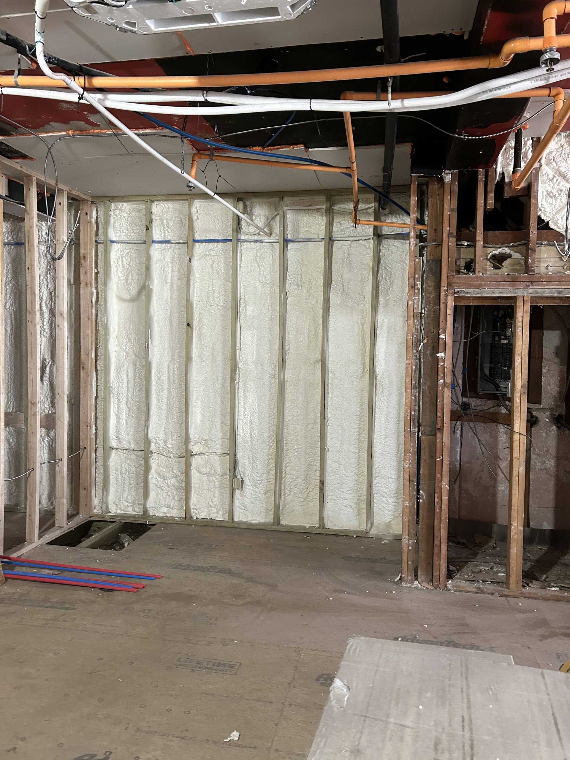 A room under renovation showing exposed wooden wall studs, spray foam insulation, and open ceiling pipes.