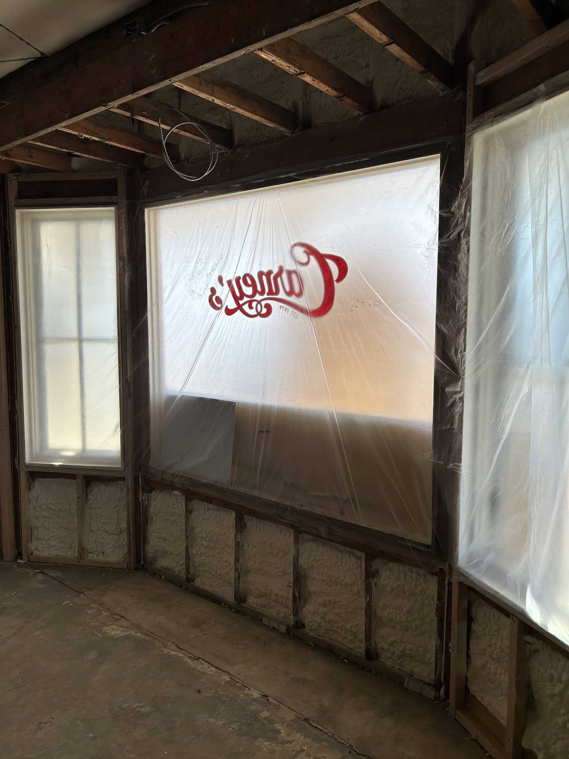 Construction site interior with exposed wall studs, insulation, and plastic sheeting covering windows.