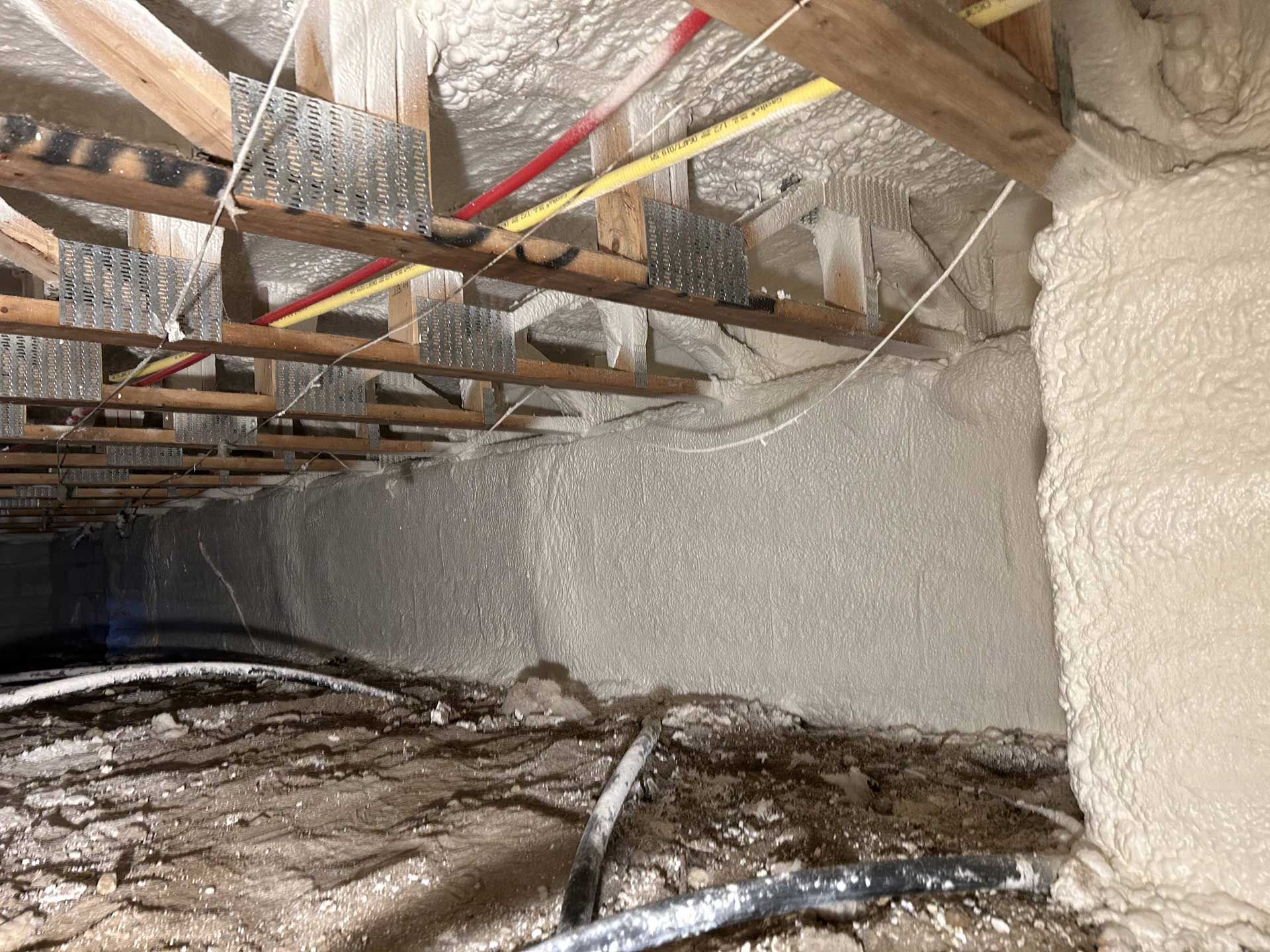 The underside of floor joists with metal connector plates, spray foam insulation on walls, and debris on the crawlspace floor.