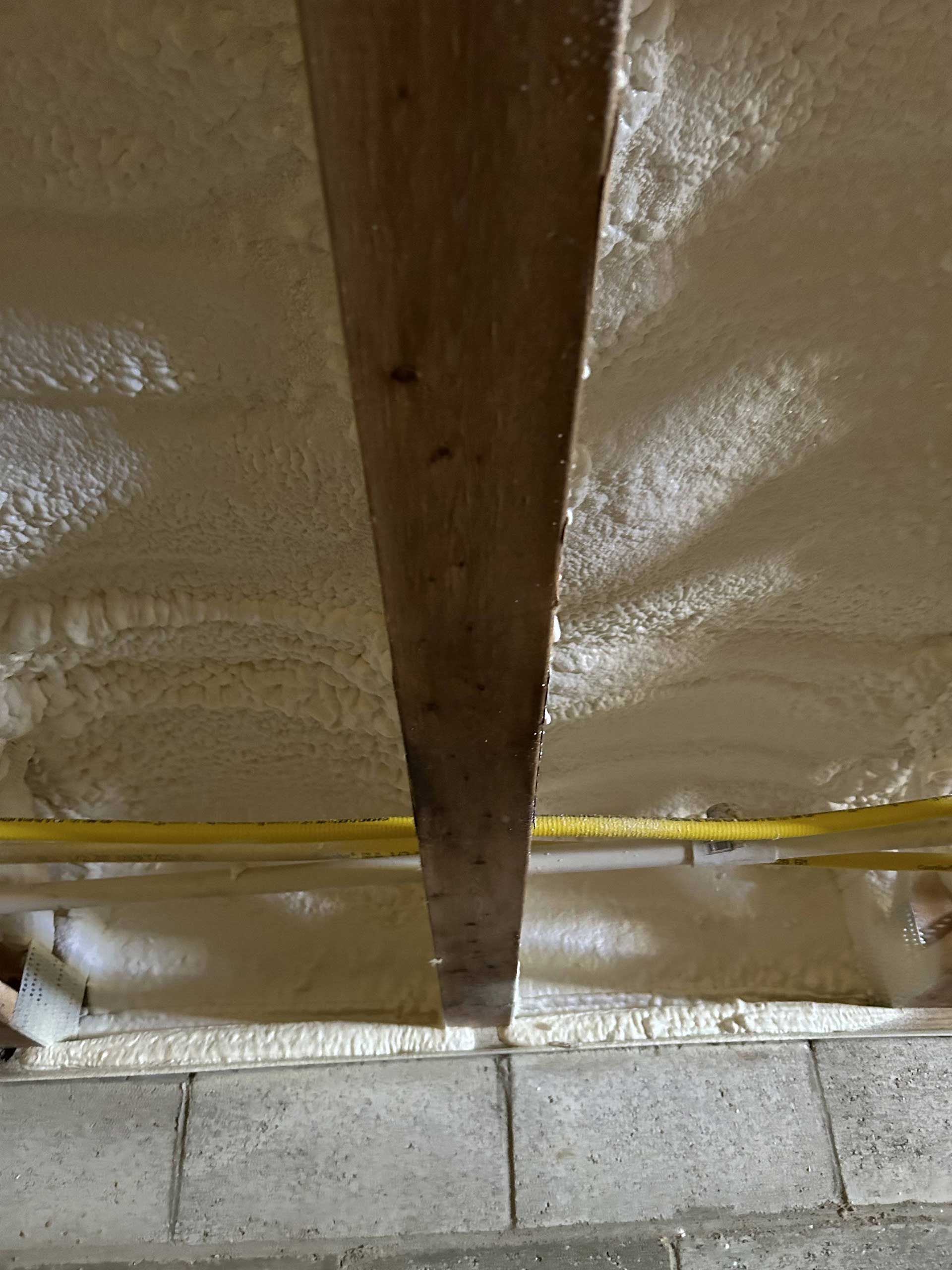 Spray foam insulation applied to the underside of a roof between wooden rafters, with yellow pipes running horizontally.