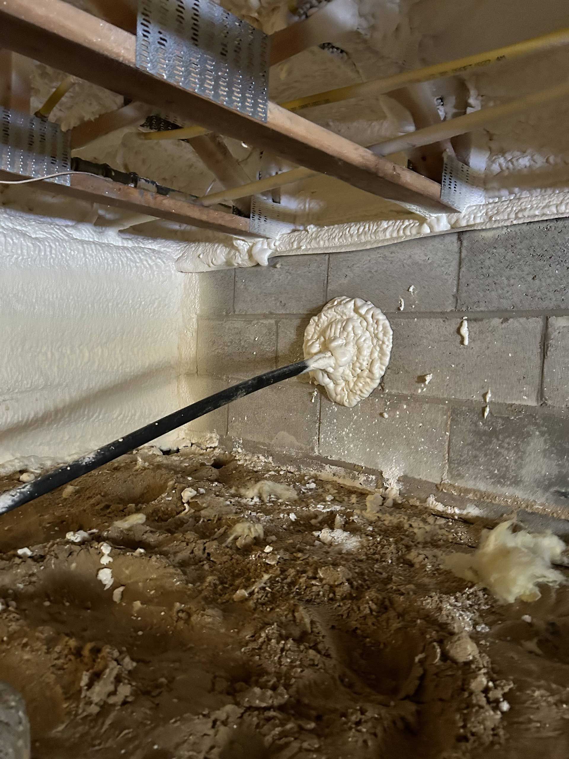 A pipe enters a crawl space through a concrete block wall, sealed with expanding foam, beneath floor joists with insulation.