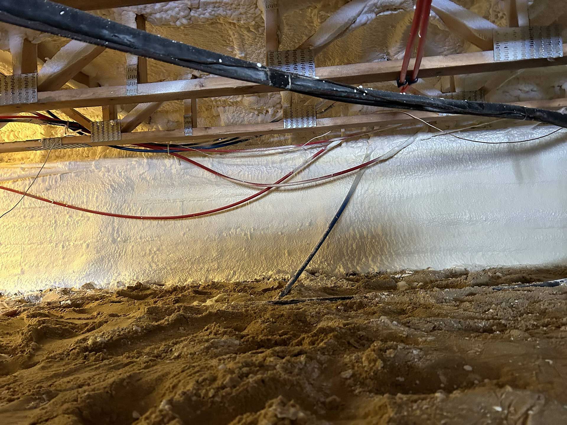 Spray foam insulation applied to the foundation walls and ceiling joists in a dirt-floored crawlspace with exposed wiring.