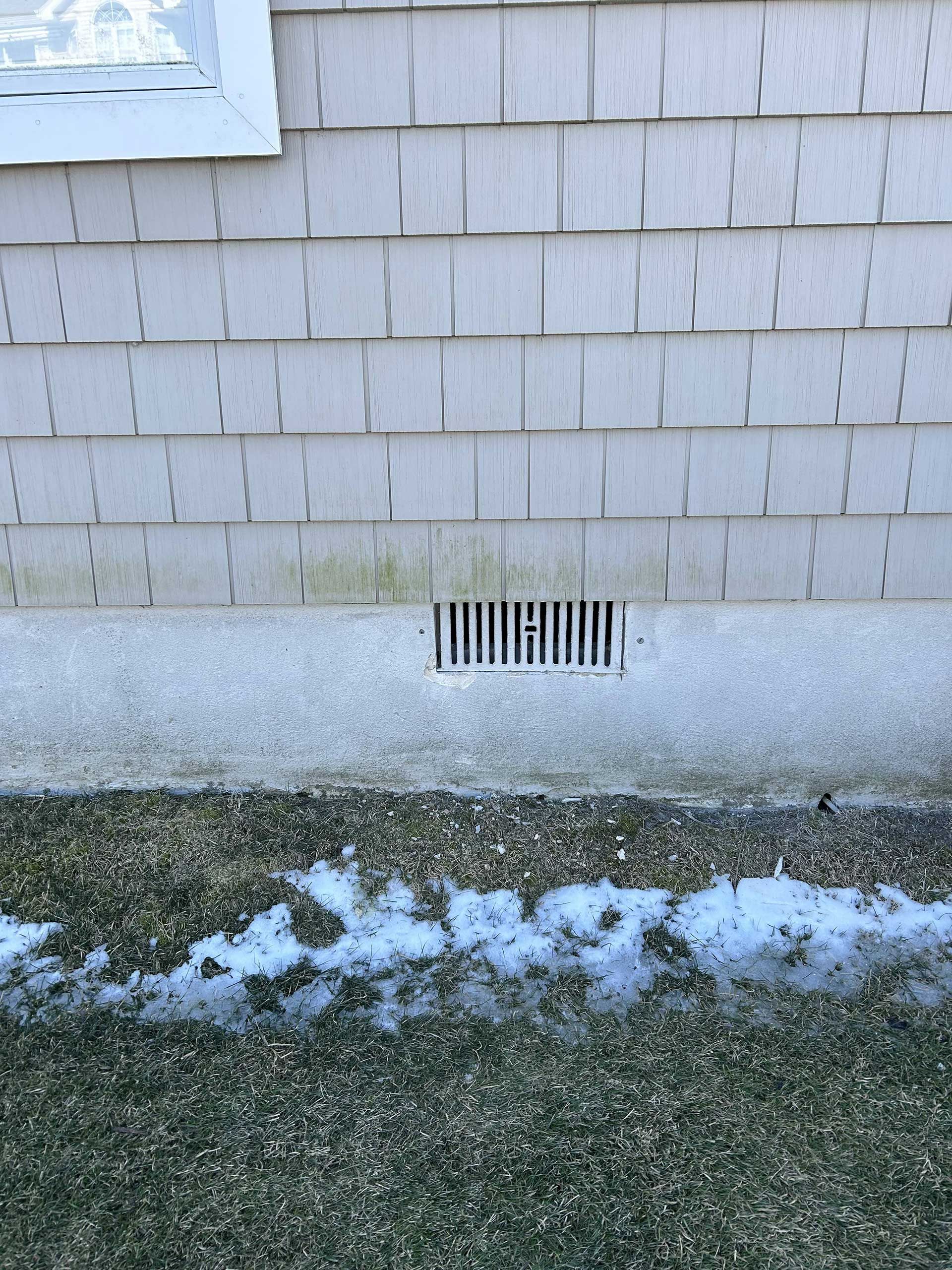 A vent in a concrete house foundation below wood shingles, with light green mold and snow-covered ground in front.