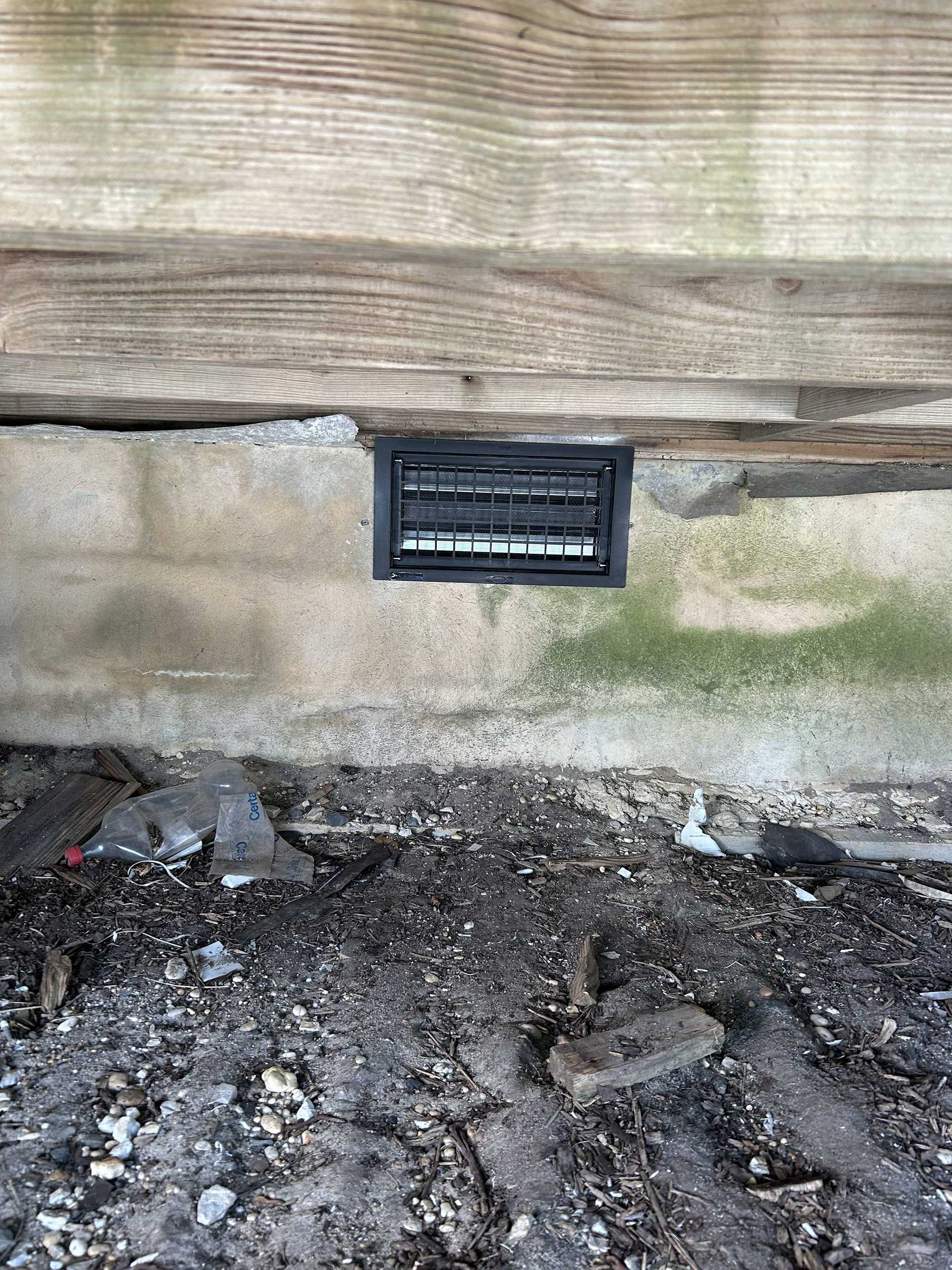 A black rectangular crawl space vent embedded in a concrete foundation wall beneath a wood sill plate.