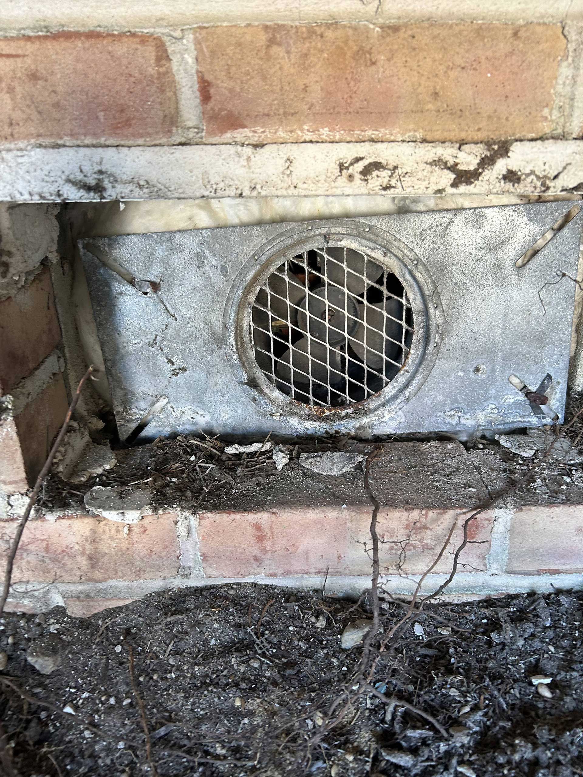 A metal rectangular vent cover with a circular grille installed in a brick foundation wall above a dirt-covered surface.