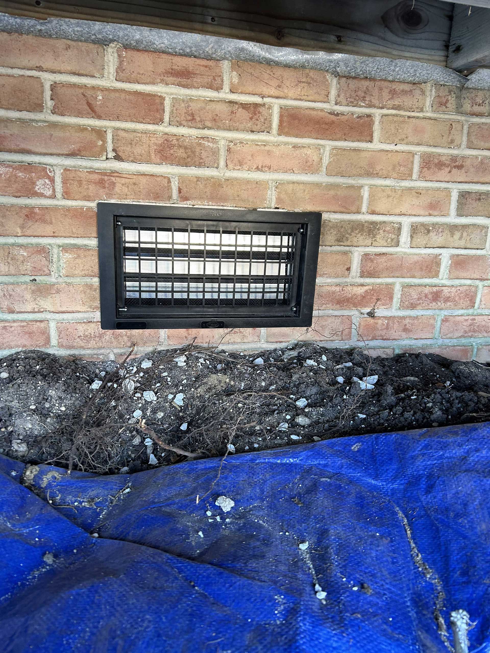 A black vent in a brick wall, situated above a pile of dark soil and a blue tarp.