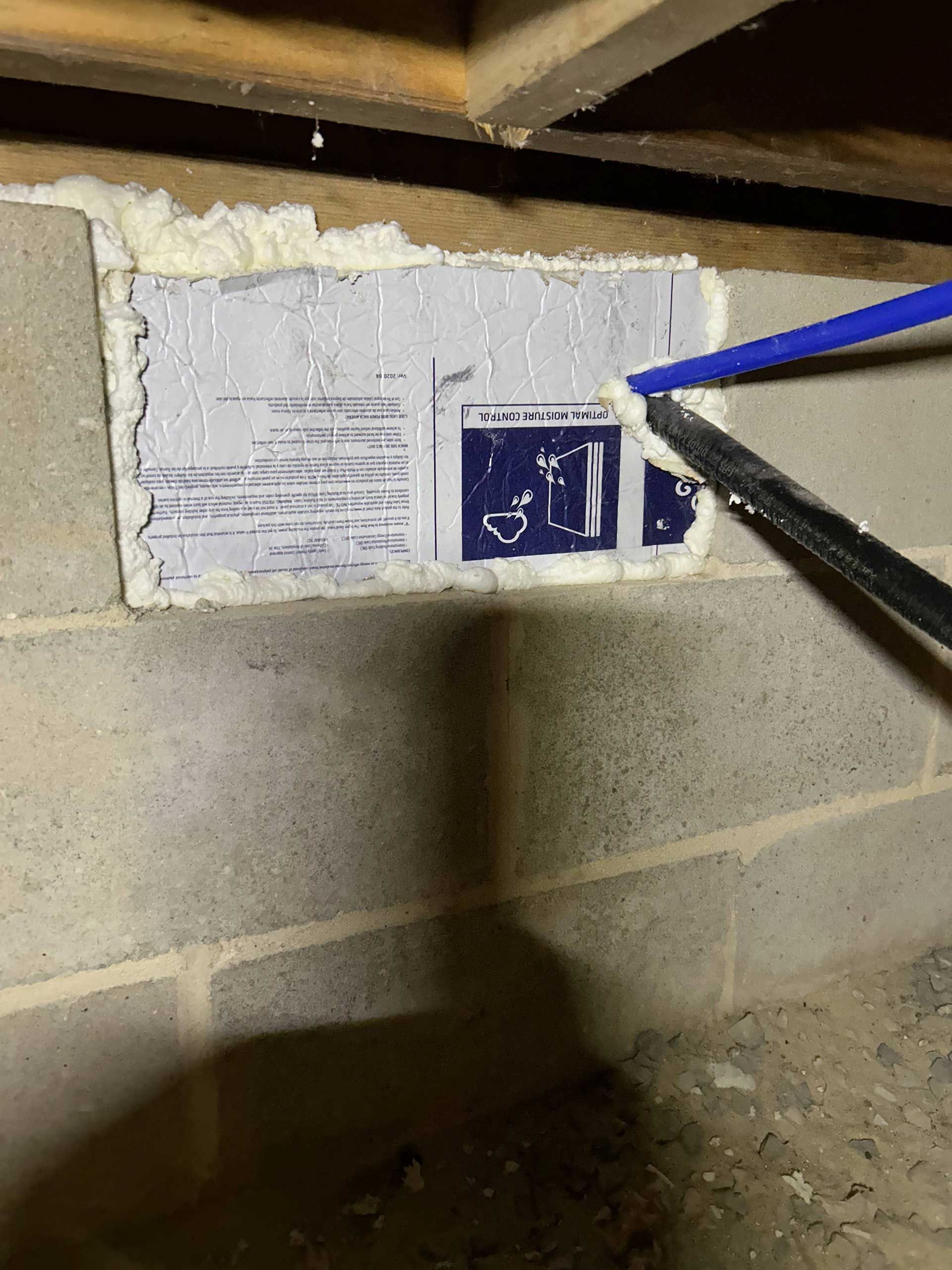 A piece of foam board insulation sealed with spray foam into a crawl space foundation vent, pointed to by a blue tool.