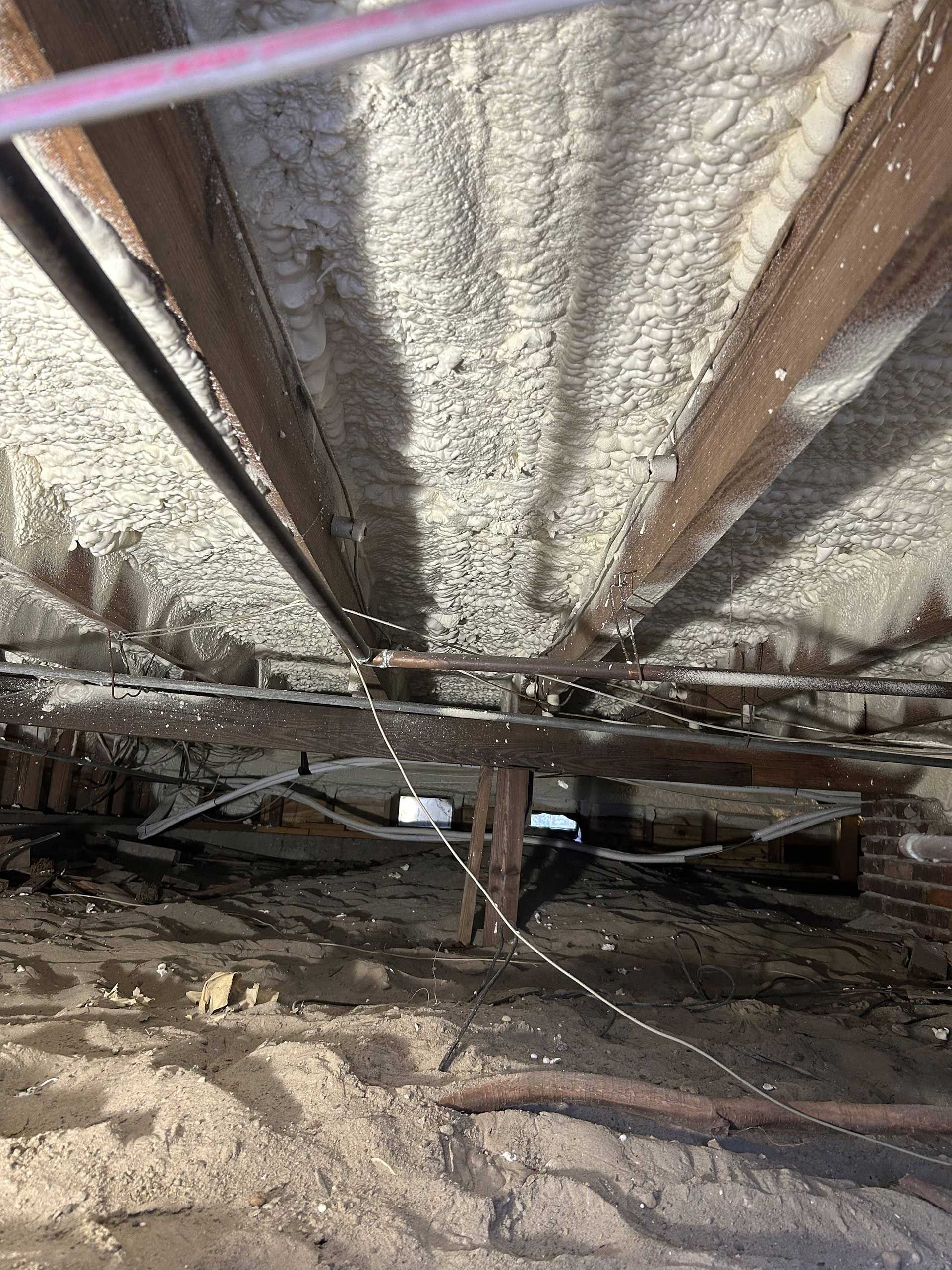 Foam insulation applied between floor joists above a crawl space with exposed pipes and dirt ground.