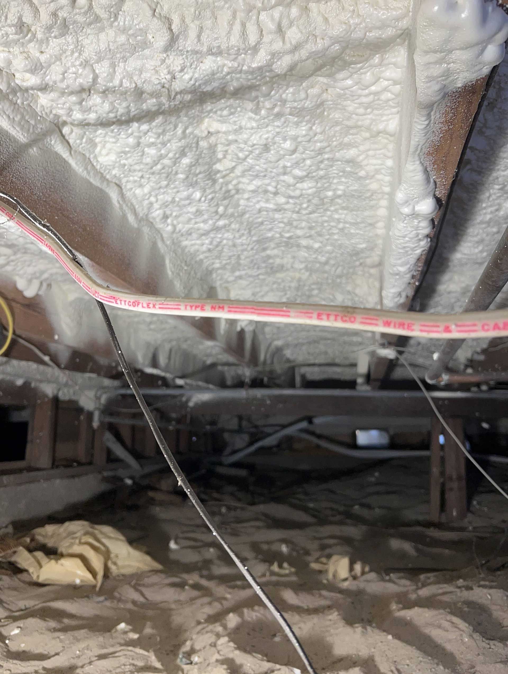 A crawl space with spray foam insulation applied between floor joists above a dirt ground with exposed wires and pipes.