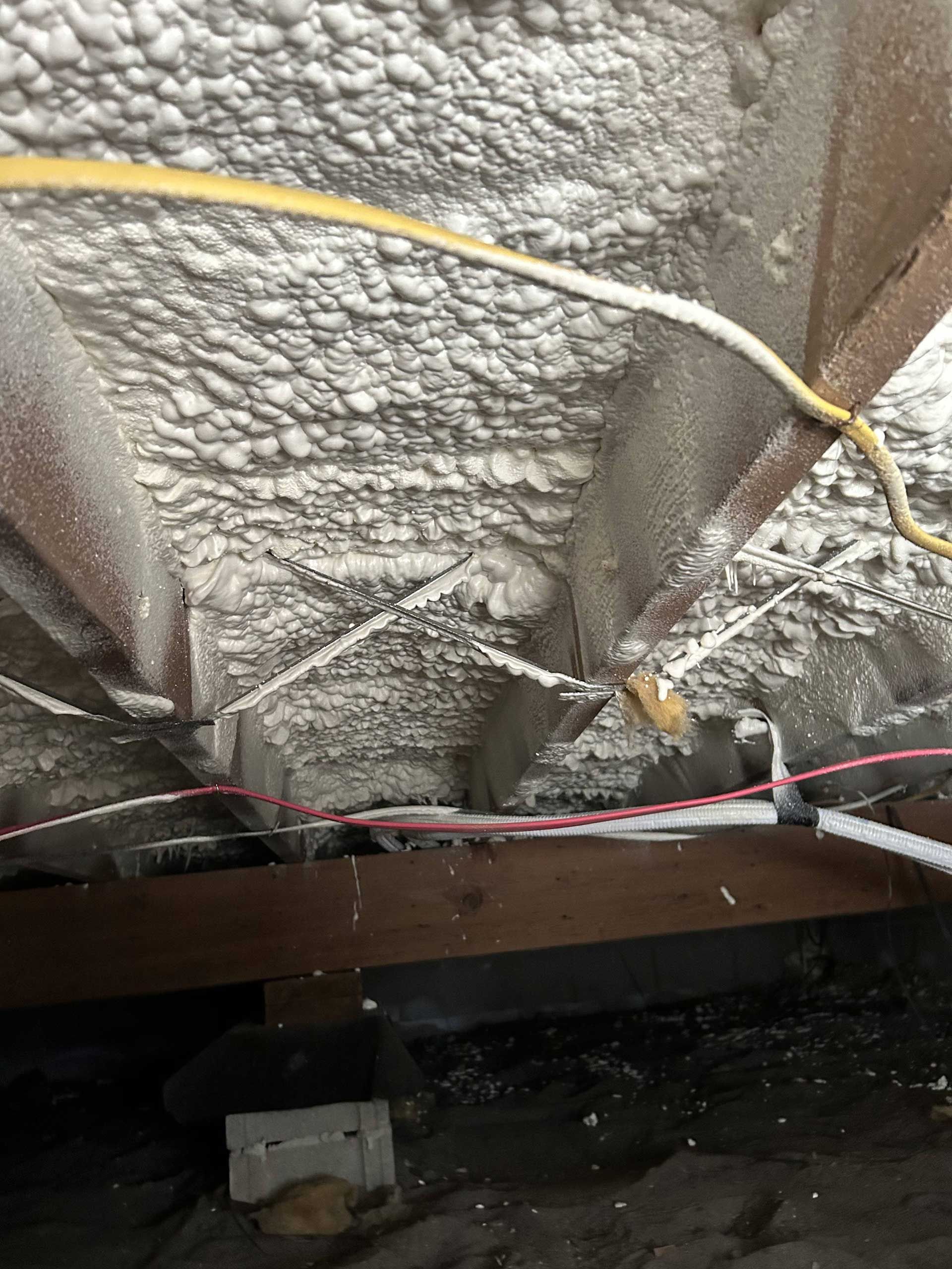 Spray foam insulation applied to the underside of a floor, with visible electrical wiring and wooden joists in a crawlspace.