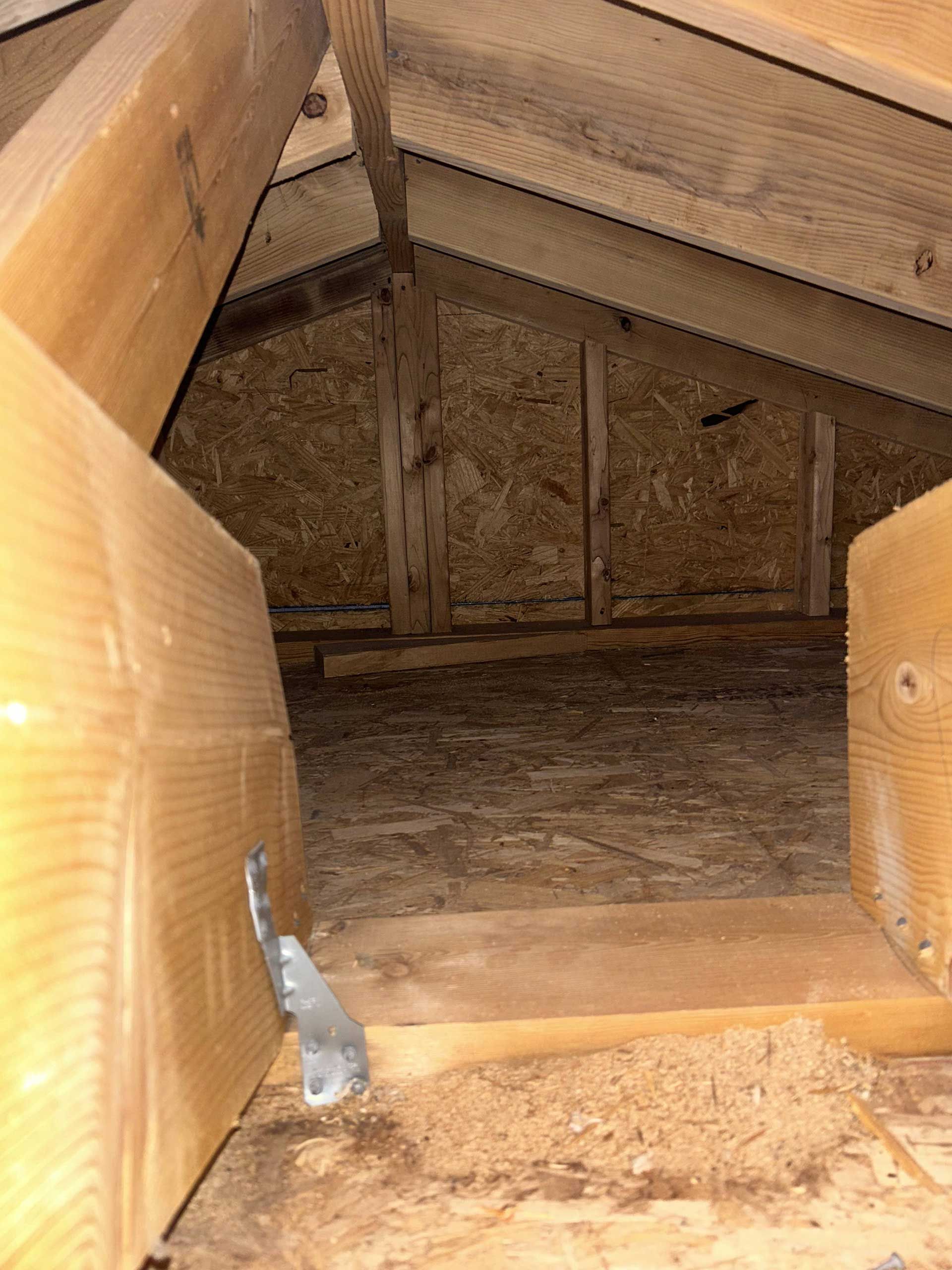 A view inside a wooden attic framing, showing rafters, floor sheathing, and a metal hurricane tie on a support beam.