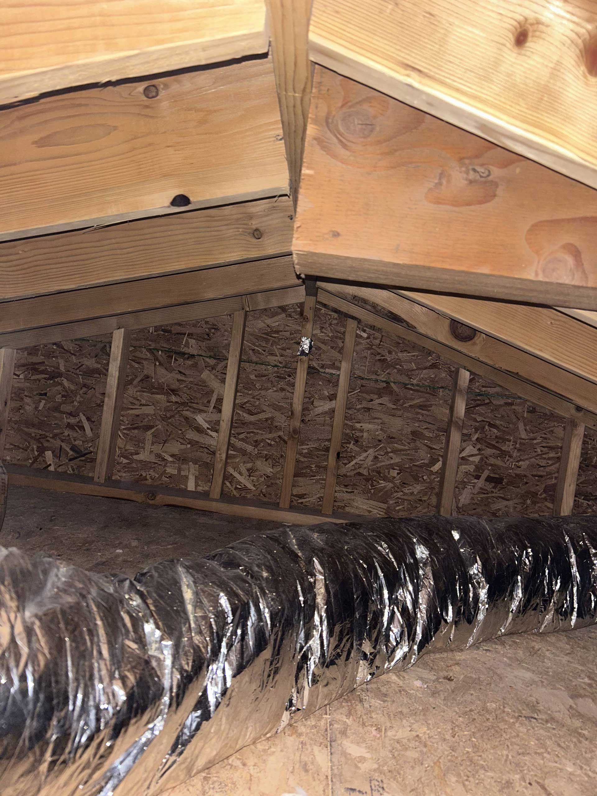 An attic interior showing exposed wooden rafters, a dark flexible ventilation duct, and fiberboard sheathing.