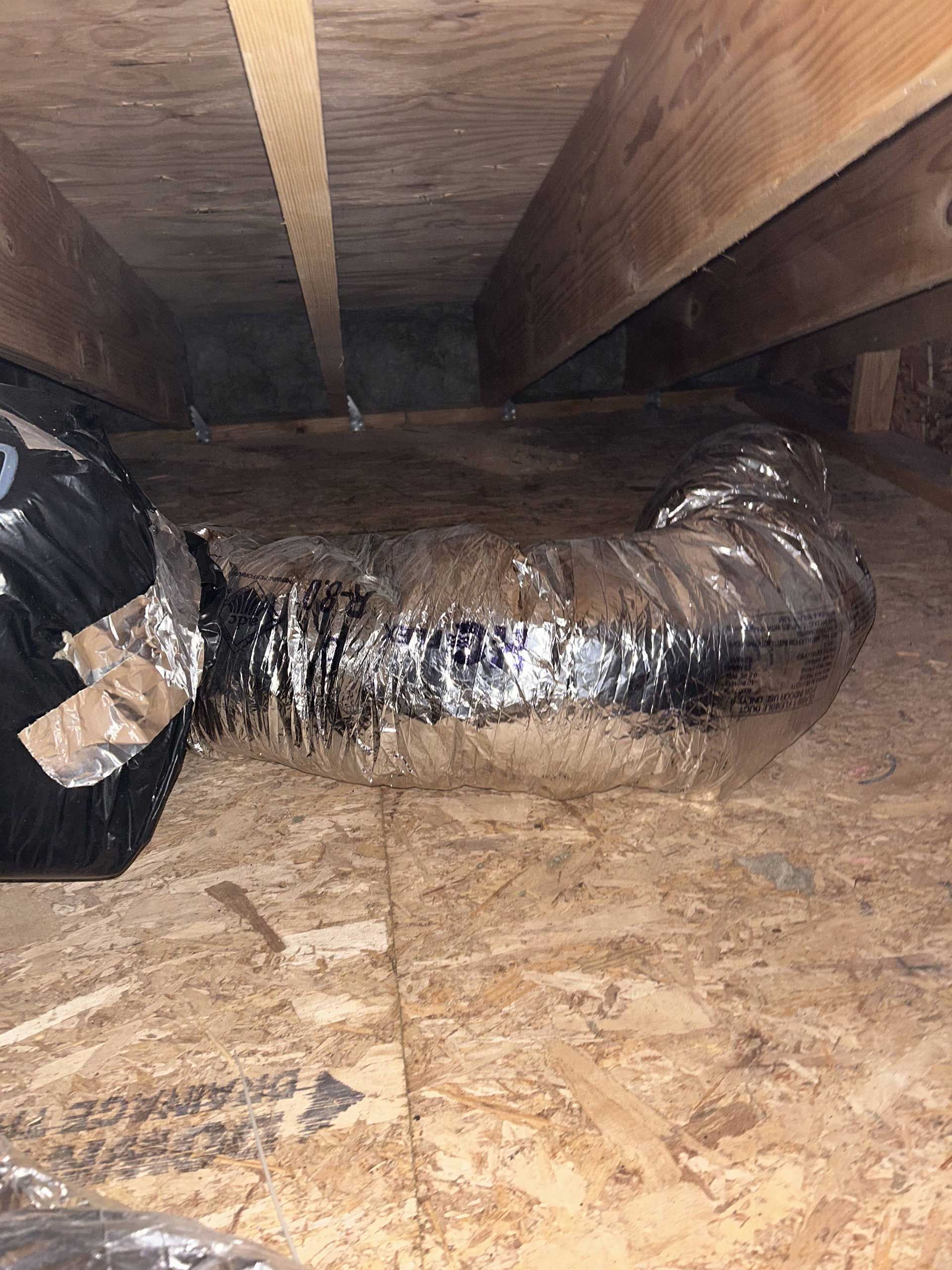 An attic space showing a flexible, silver foil HVAC duct connecting to a black junction box resting on plywood flooring.