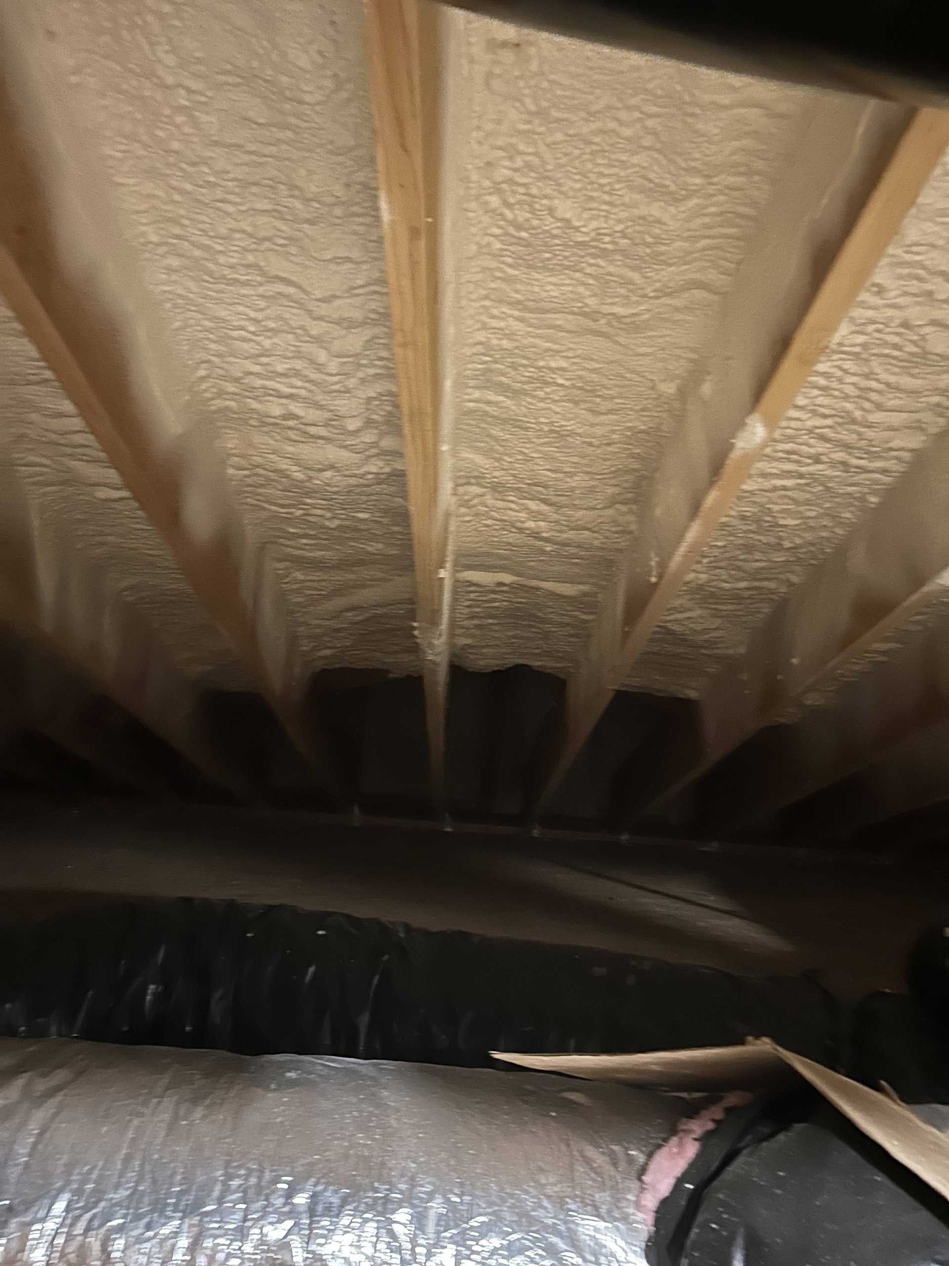 Attic rafters insulated with beige spray foam, featuring HVAC ductwork in the foreground.