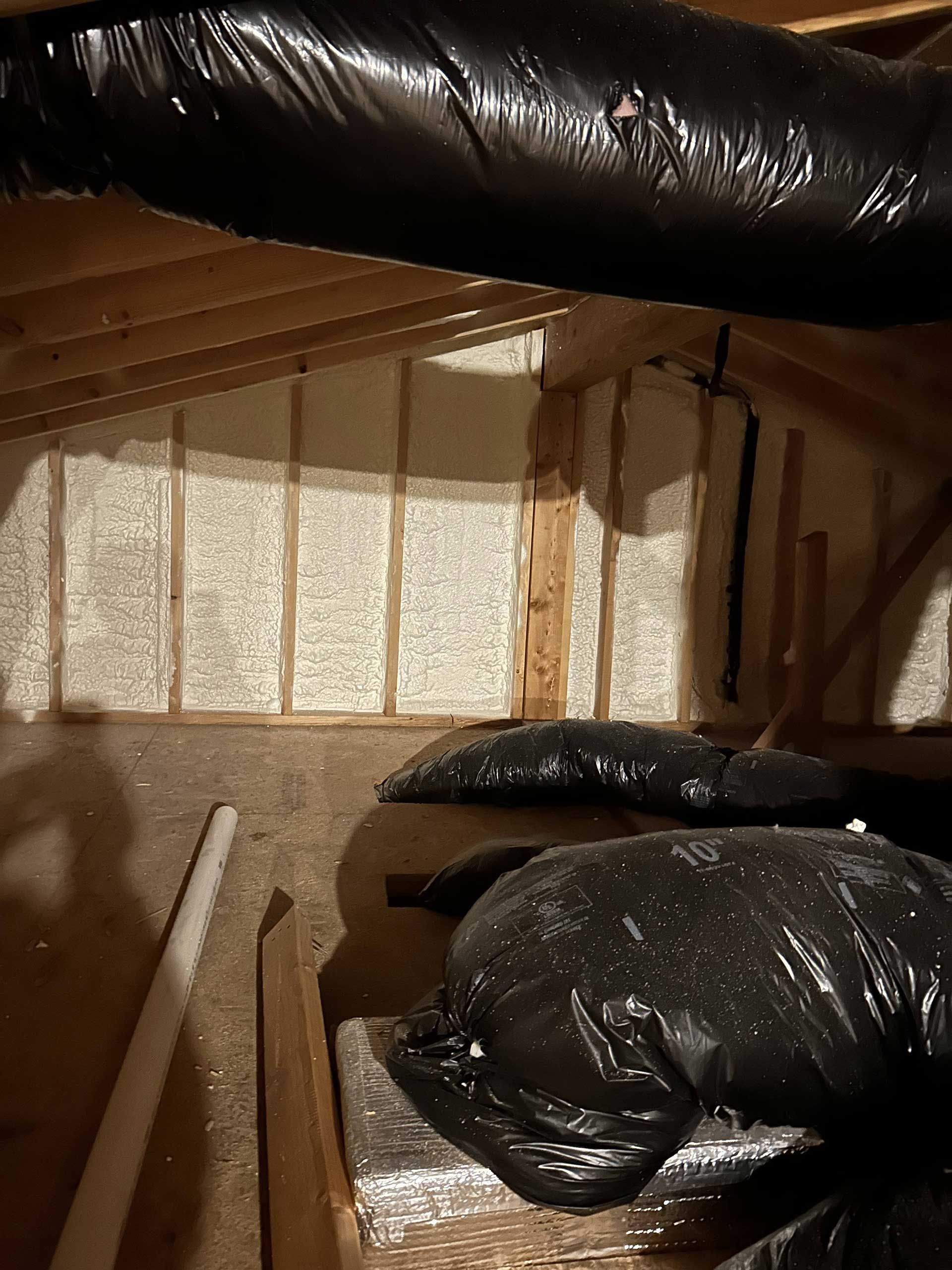 An attic space featuring a sloped roof insulated with spray foam, wooden framing, and black ductwork.