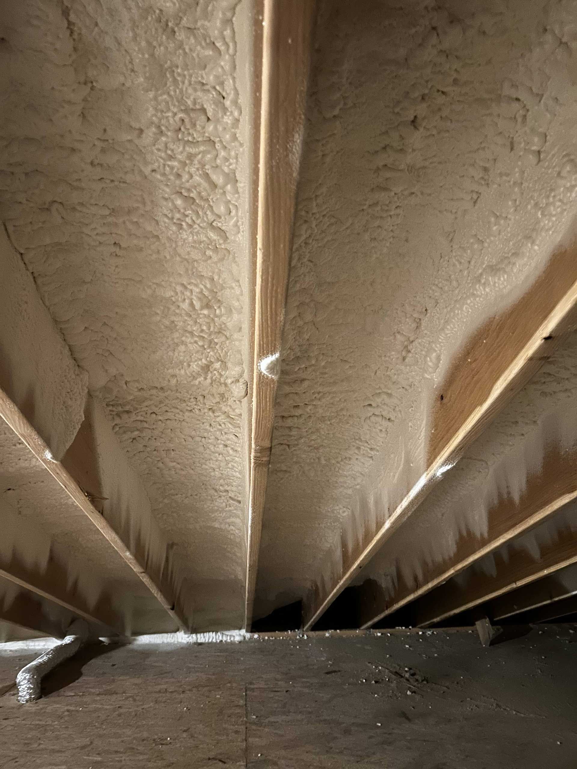 Low-angle view of wooden floor joists insulated with sprayed foam between them, viewed from a basement or attic crawlspace.