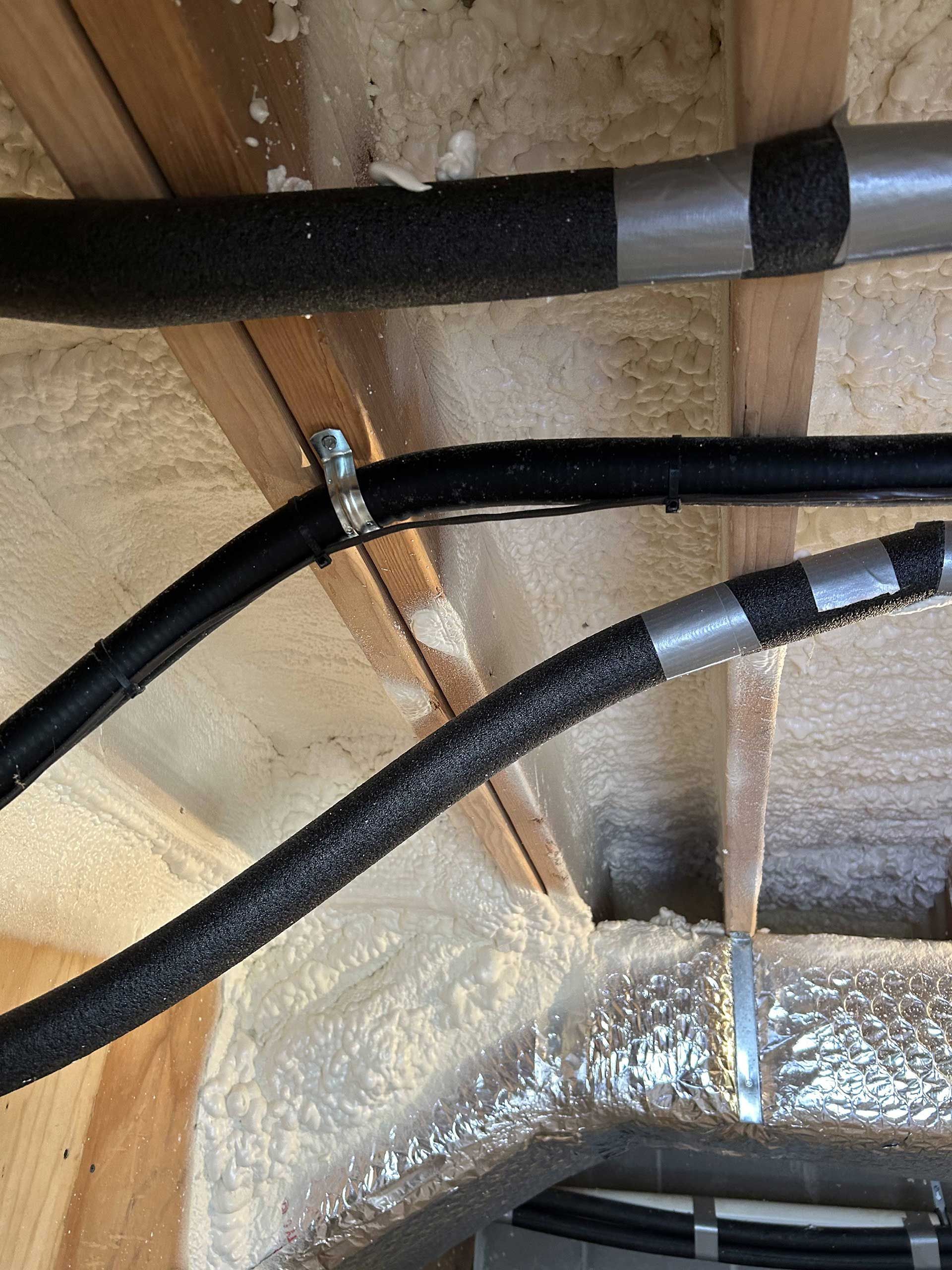 Insulated pipes run across wooden ceiling joists with spray foam insulation between them.