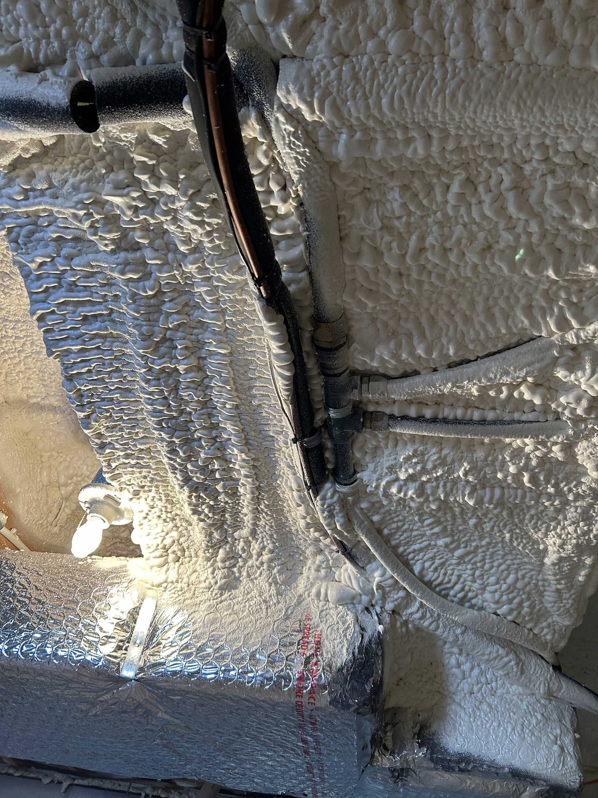 Spray foam insulation applied to a wall around a dark pipe and wires, with visible icicles forming on the surface.