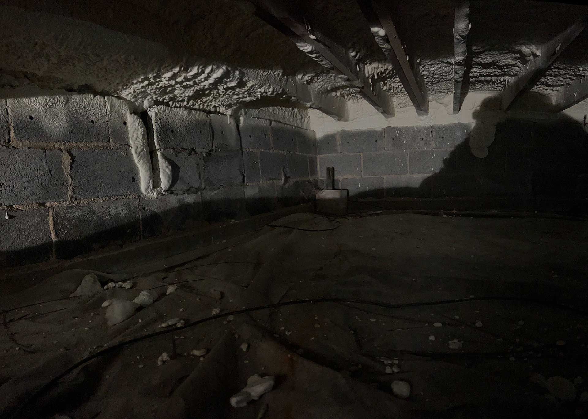 A dark crawlspace with concrete block walls, a ground vapor barrier, and white spray foam insulation on the joists.