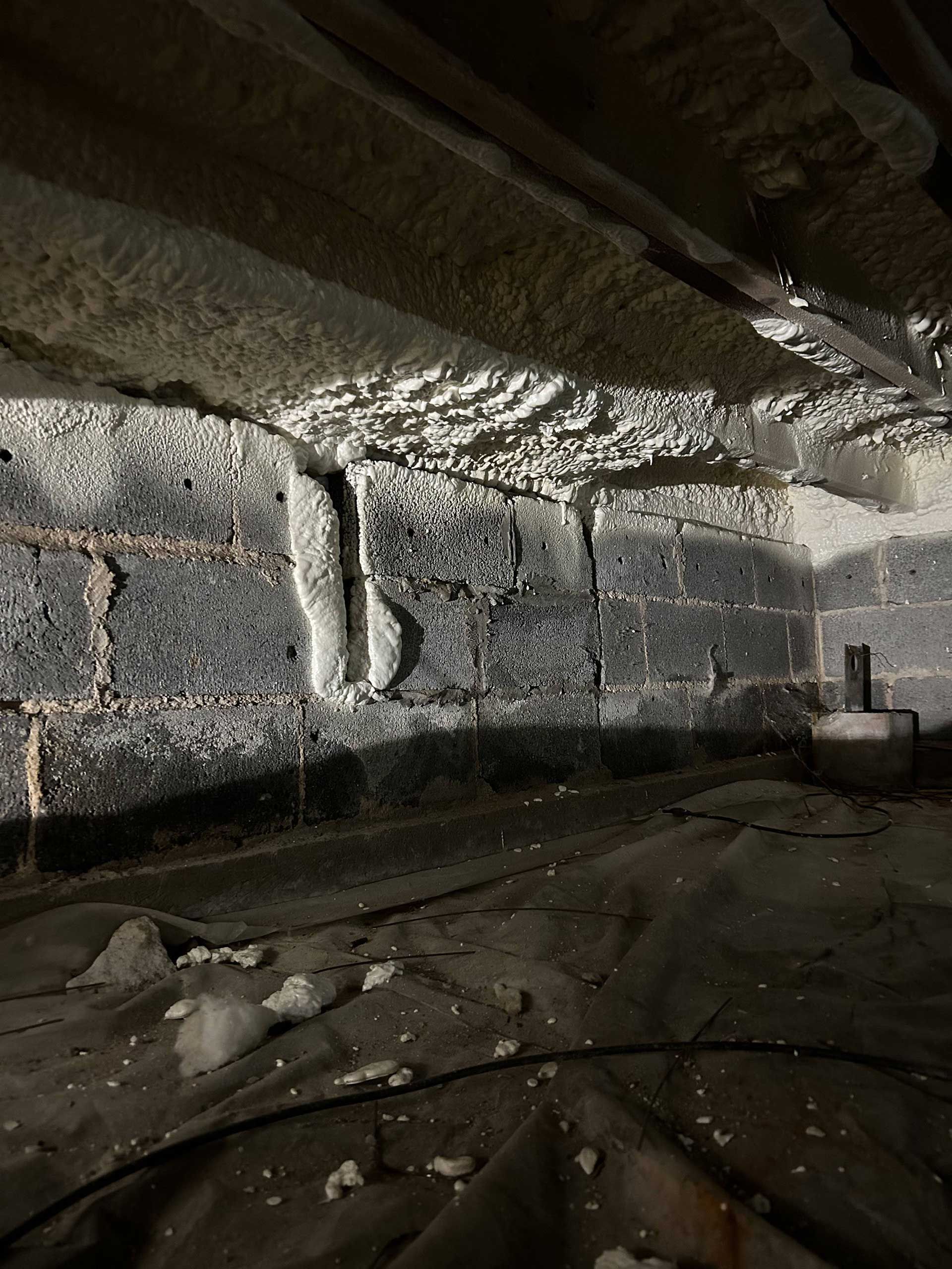 A crawl space with concrete block walls insulated with white spray foam, and a vapor barrier covering the dirt floor.