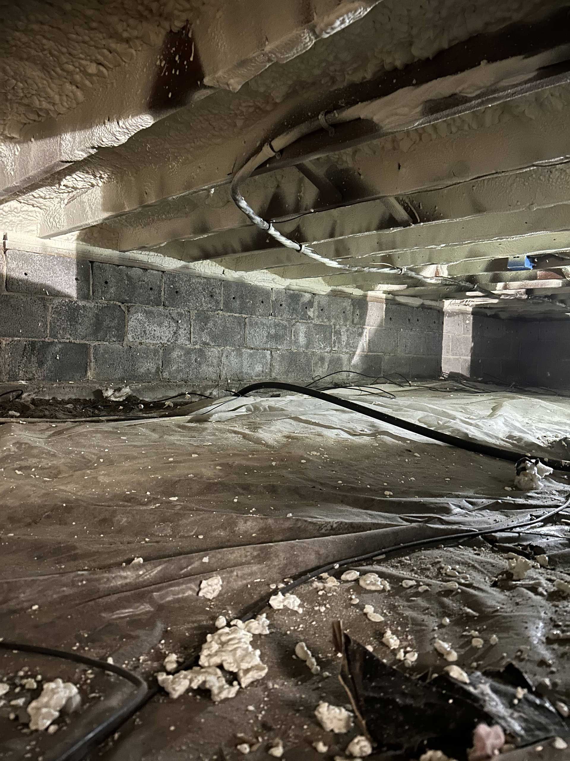 A crawlspace with spray foam insulation on ceiling joists, a dark plastic vapor barrier on the floor, and a black cable.